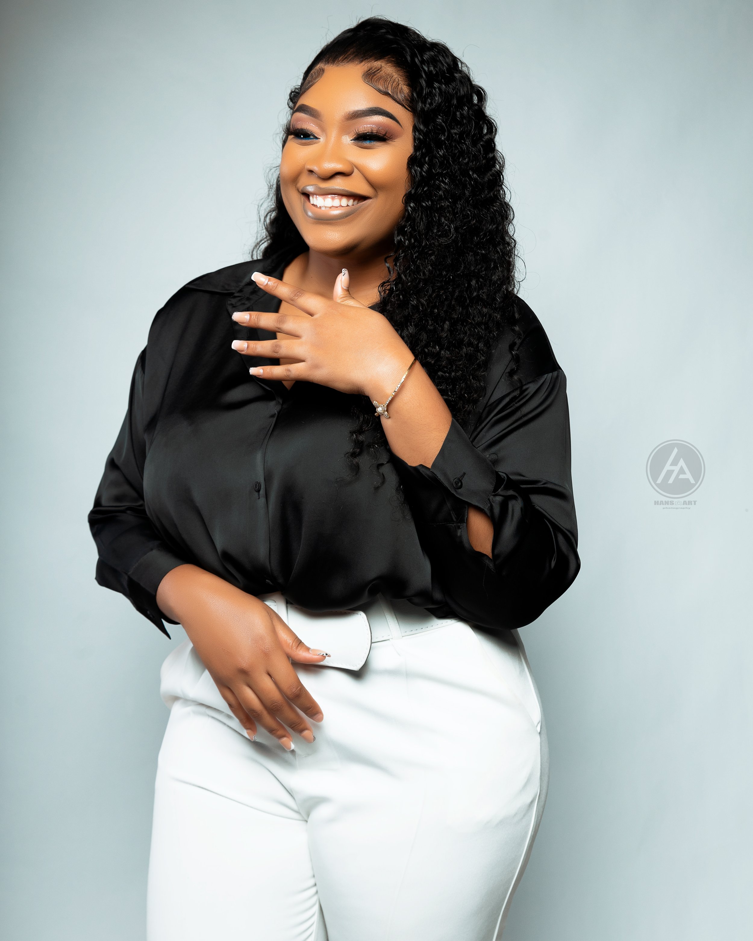 A woman with curly black hair, smiling, wearing a black satin blouse with rolled-up sleeves and white pants, standing against a plain light blue background.