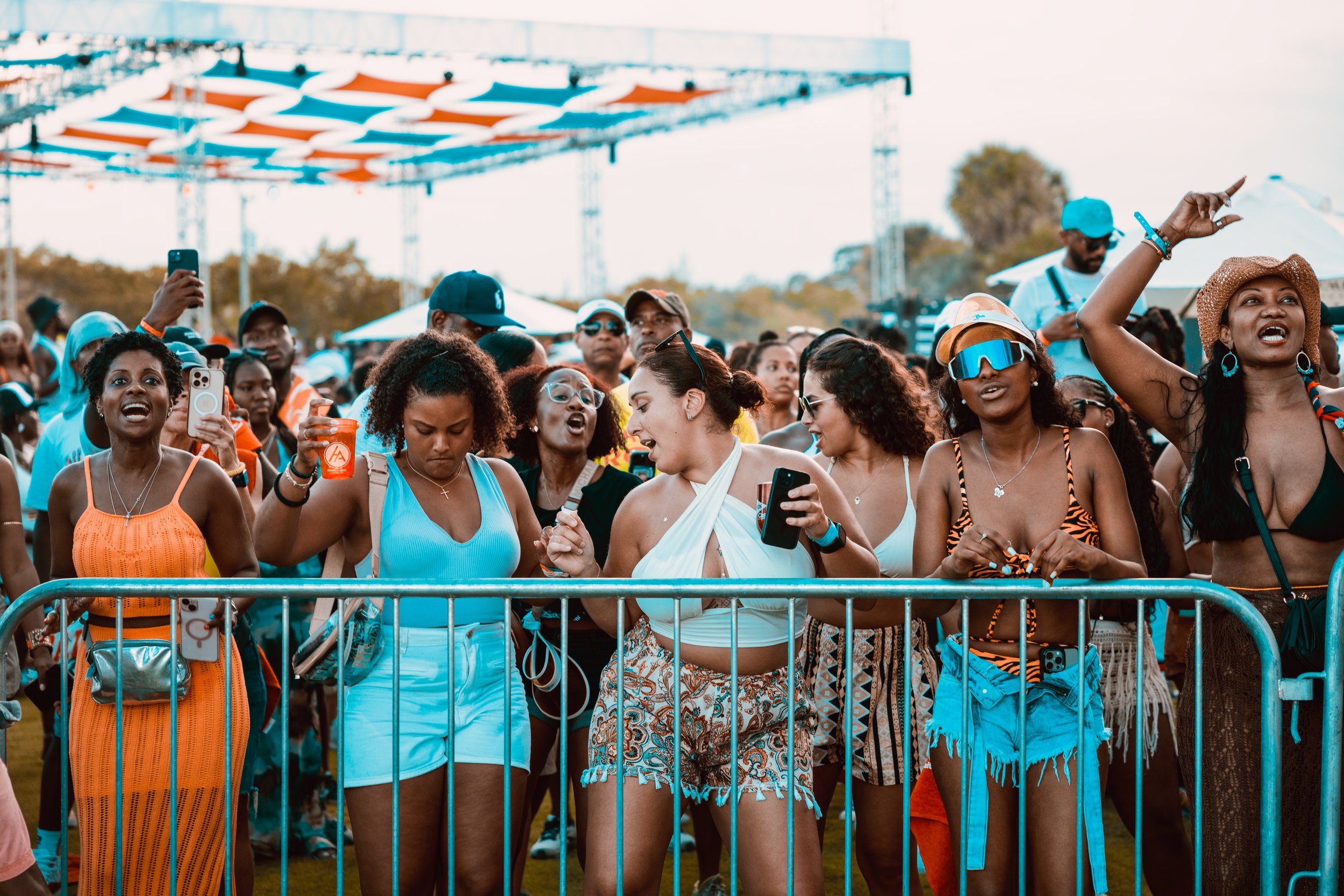 A group of women and men at an outdoor concert or festival, standing behind a barrier, some taking photos or videos with their phones, dressed in summer attire with hats and sunglasses.