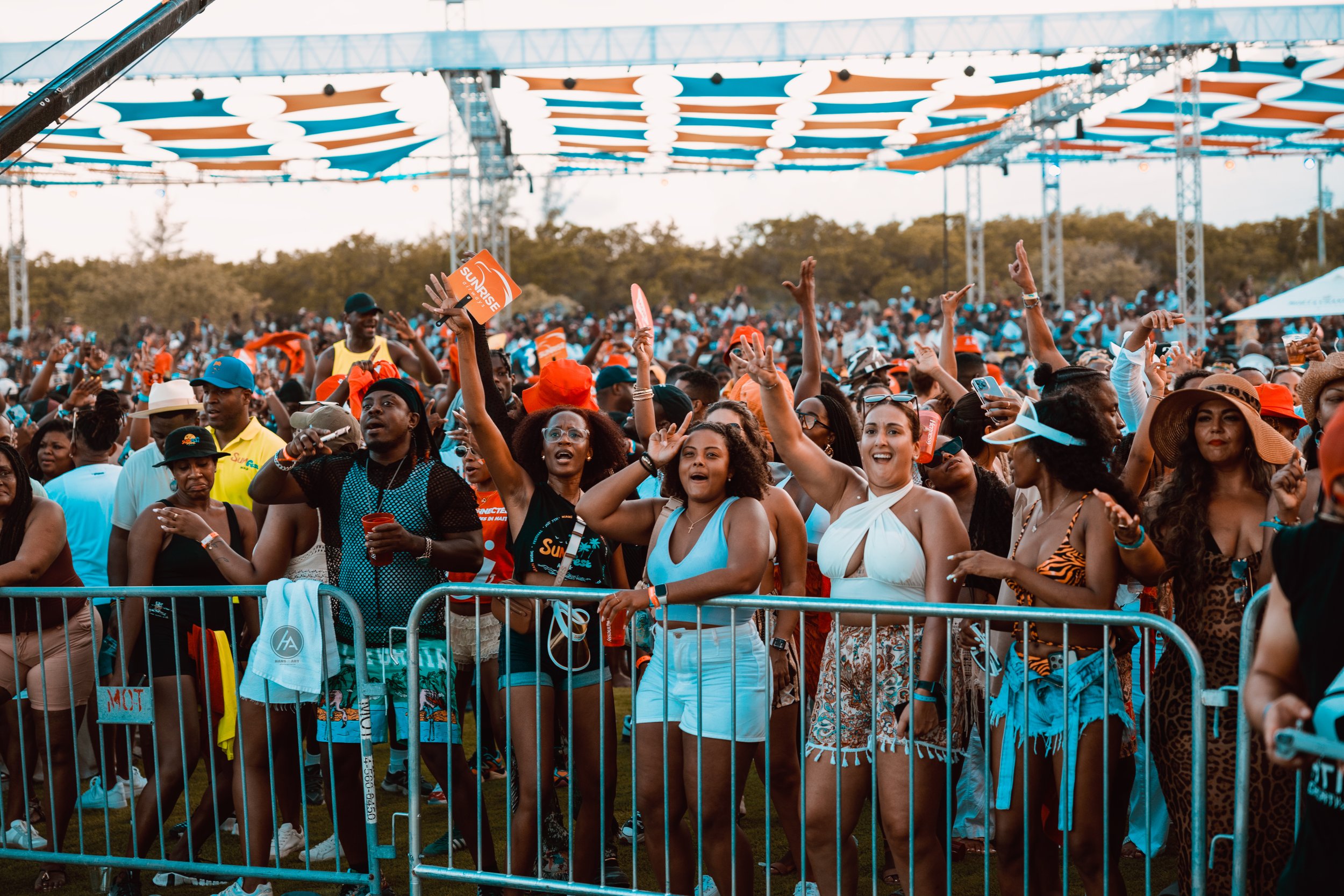 Crowd of people at an outdoor event, many wearing sunglasses and summer clothing, some raising their hands, behind a railing with a large canopy overhead.