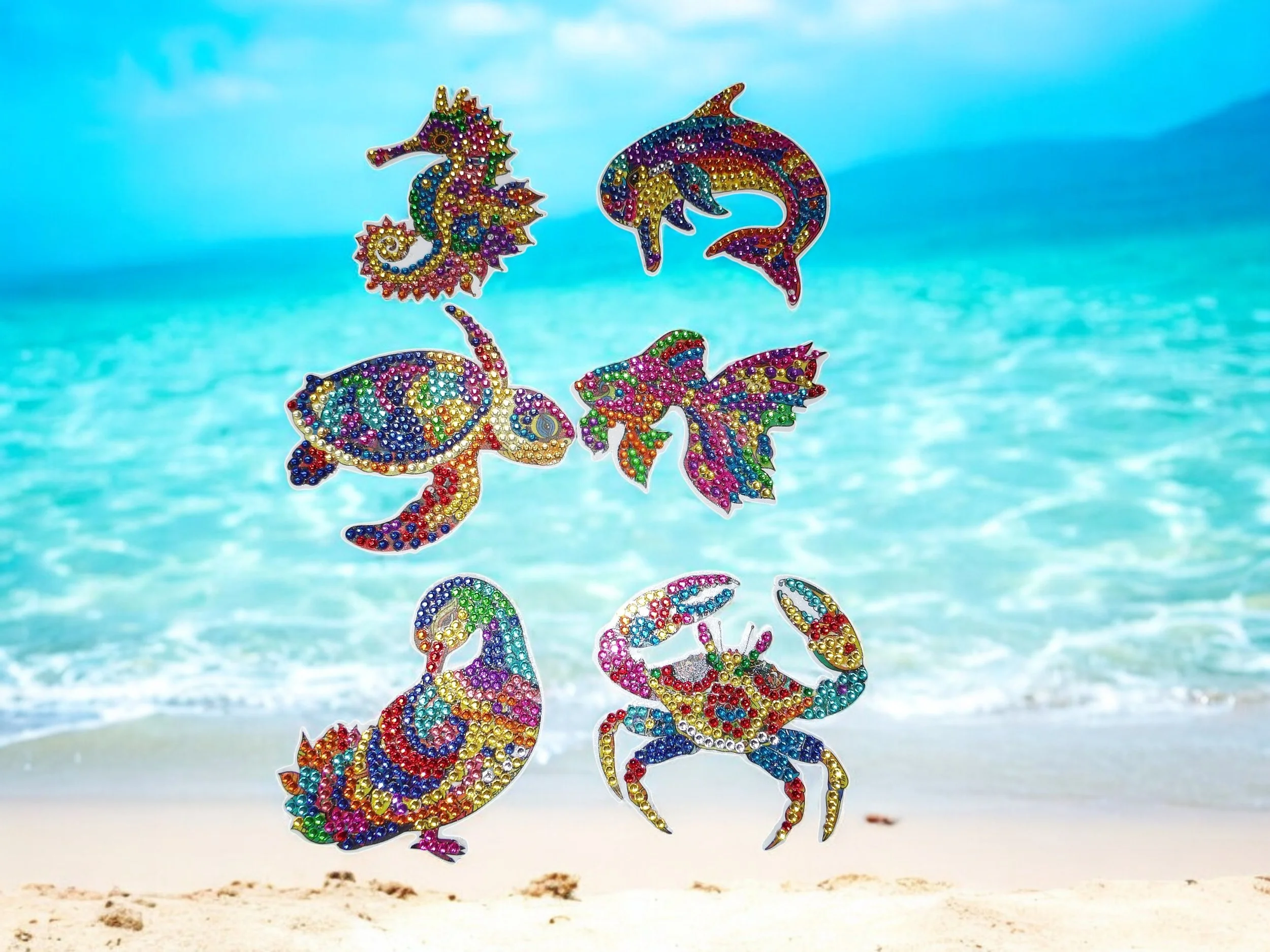 Colorful rhinestone decorations shaped like various sea creatures, including seahorses, a dolphin, a turtle, a fish, a mermaid, and a crab, hanging in front of a beach scene with sand, ocean, and sky.