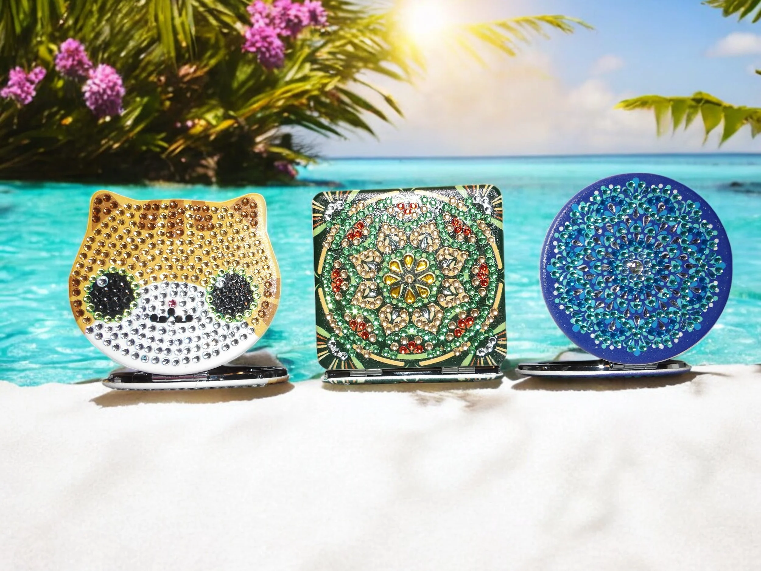 Three decorated portable mirrors with rhinestone embellishments on a beach with turquoise water, tropical plants, and a blue sky in the background.