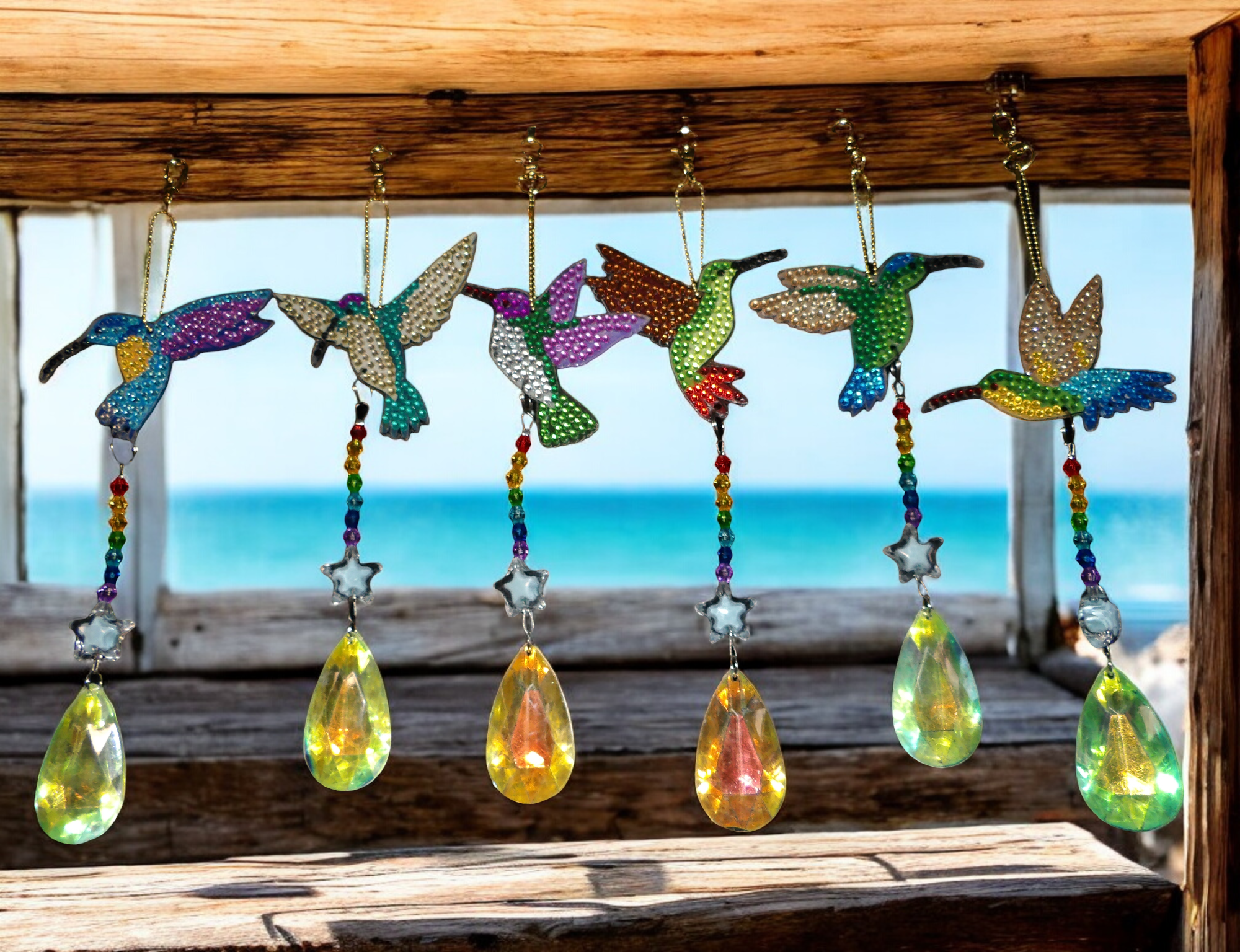 Colorful hummingbird wind chimes hanging on a wooden frame with the ocean in the background.