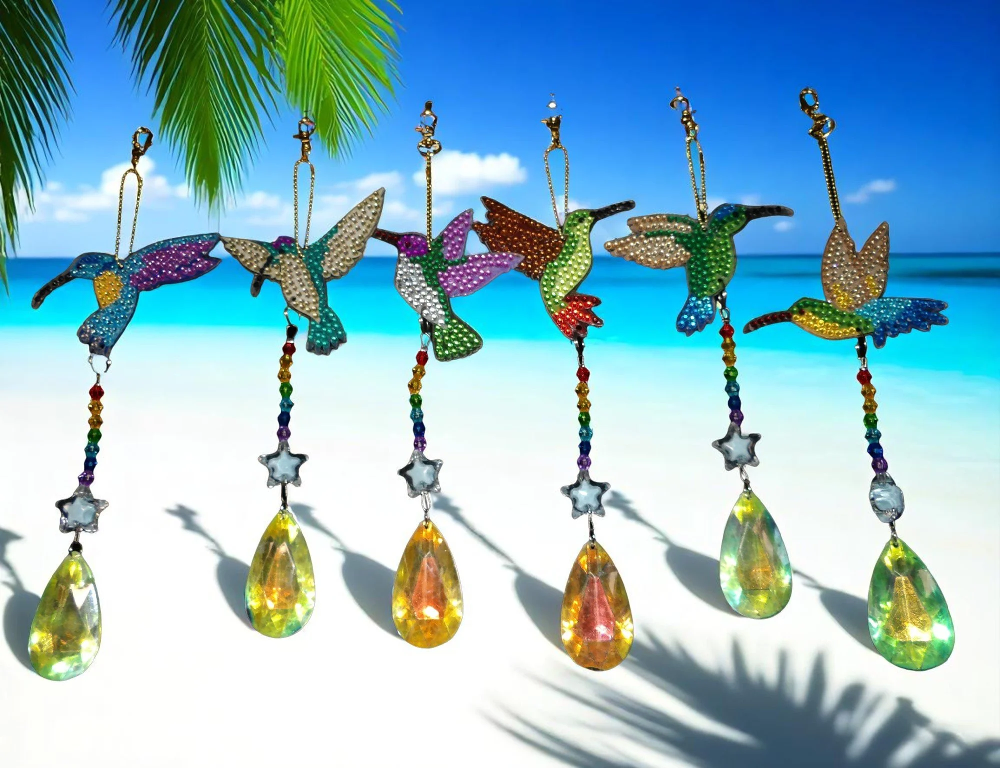 Colorful beaded hummingbird wind chimes hanging against a beach background with blue sky, ocean, and palm leaves.
