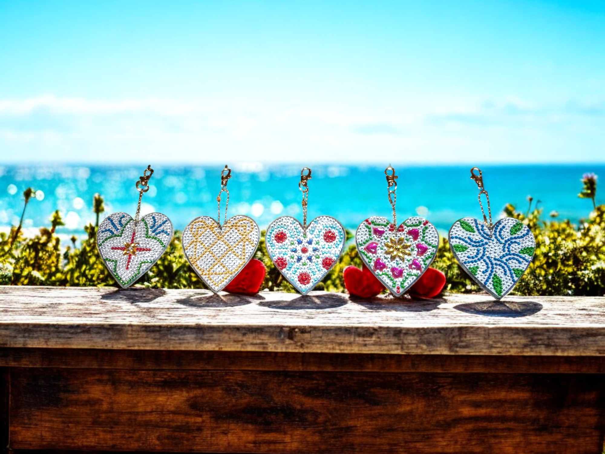 Five heart-shaped decorative keychains with colorful patterns, placed on a wooden surface, with an ocean and blue sky in the background.