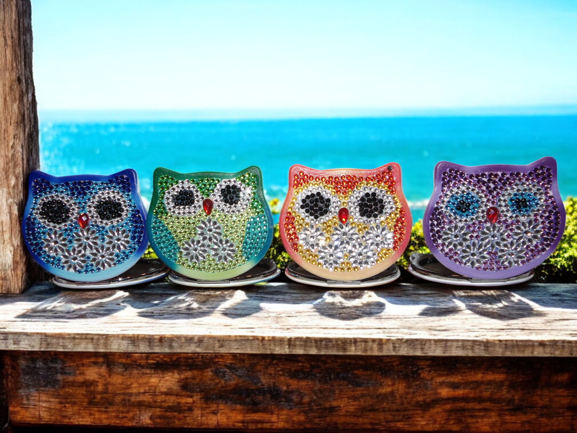 Four colorful owl-shaped candle holders decorated with rhinestones, placed on a wooden surface outdoors with a beach and ocean in the background.