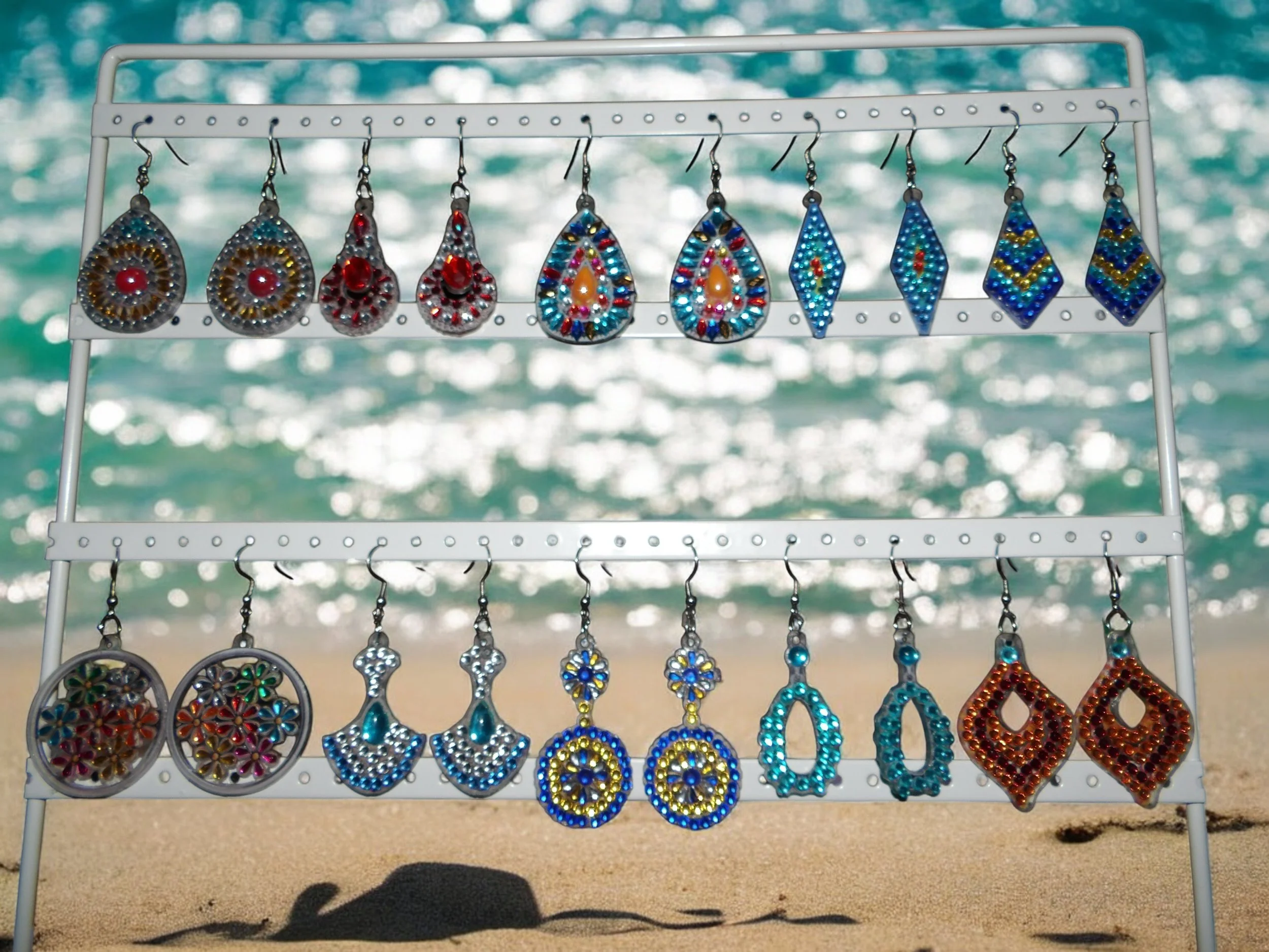 Colorful beaded earrings displayed on a white rack at the beach, with ocean waves in the background.