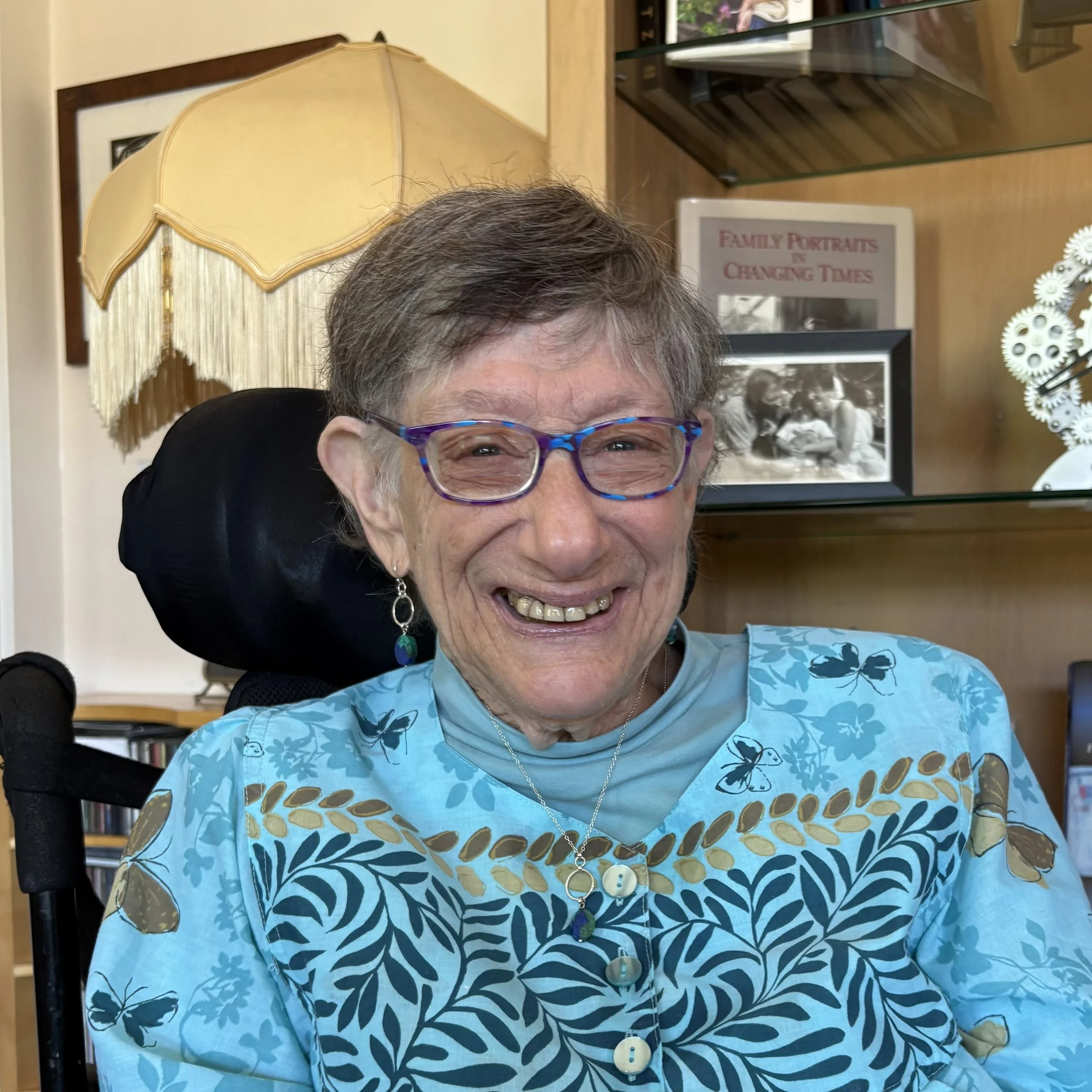 A smiling elderly woman with glasses and short gray hair, wearing a blue patterned top and jewelry, seated indoors near a lamp and a display shelf with family photos and books.