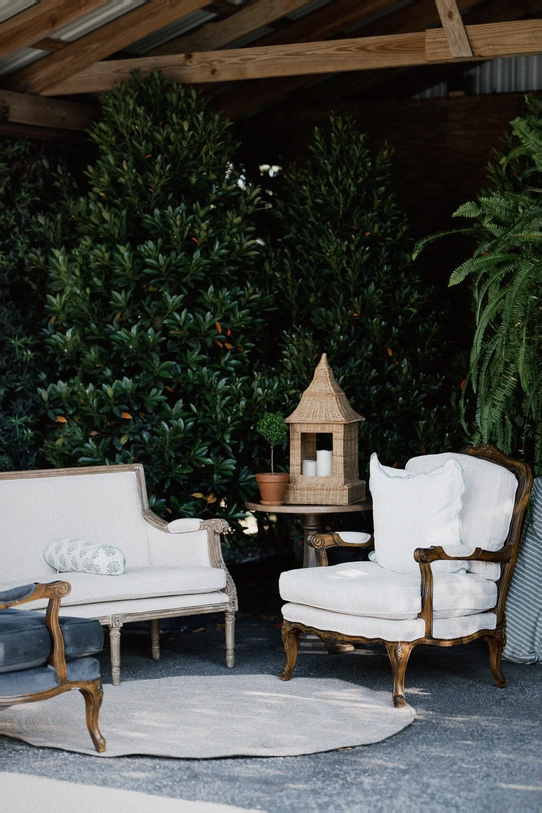 Living area with vintage white armchair, cream-colored loveseat, small side table with a potted plant and a decorative lantern, and a lush green garden background.