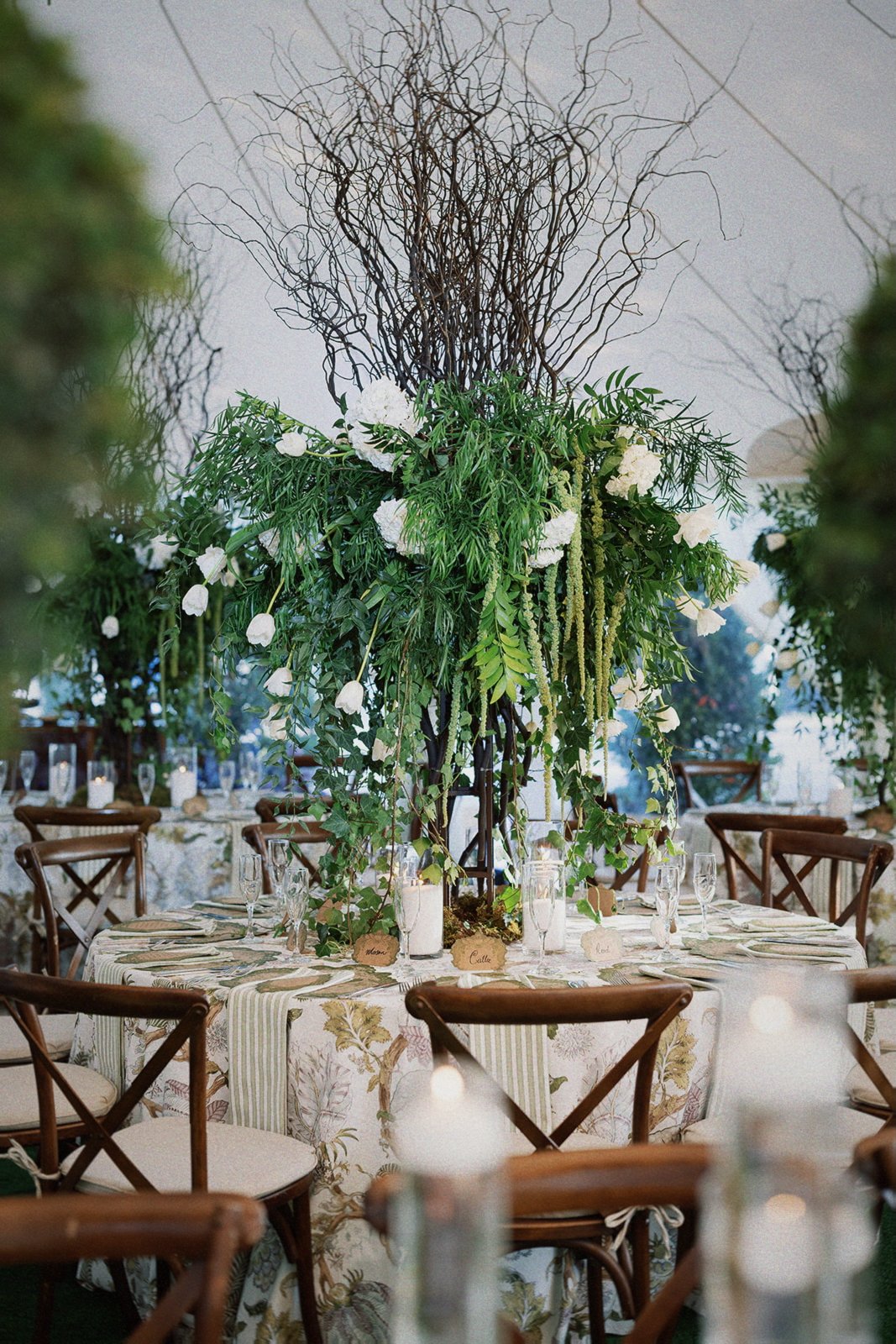 A decorated banquet table with a tall floral centerpiece consisting of green foliage, white flowers, and intertwined brown twigs in an interior event setting.