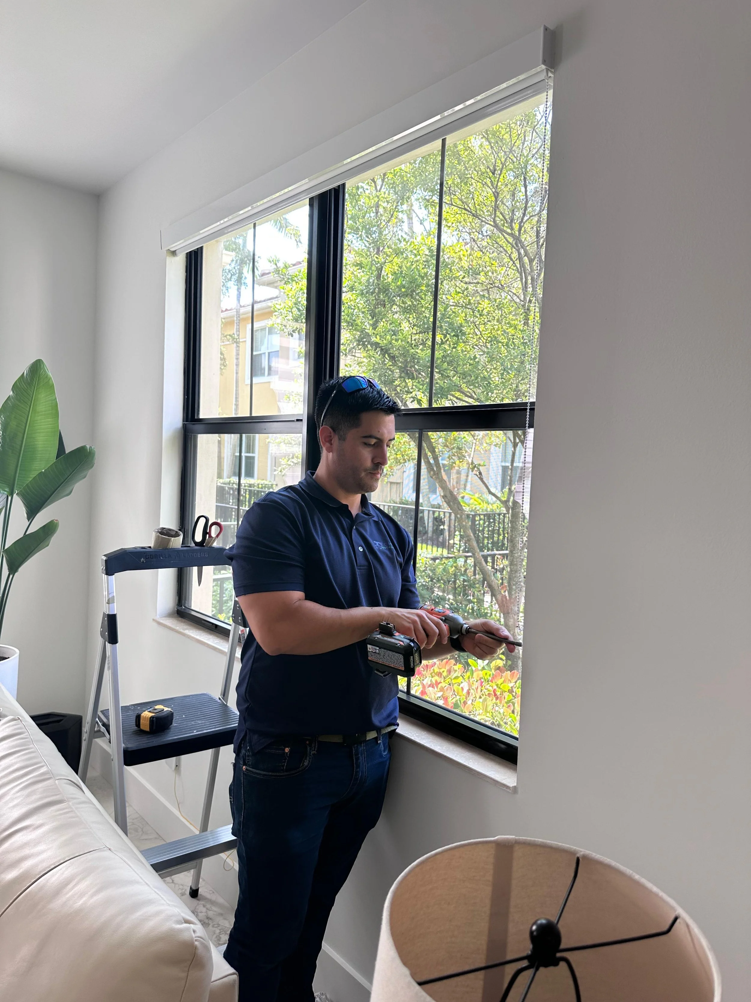 A man standing on a step ladder installing or repairing a window with a drill inside a bright living room.