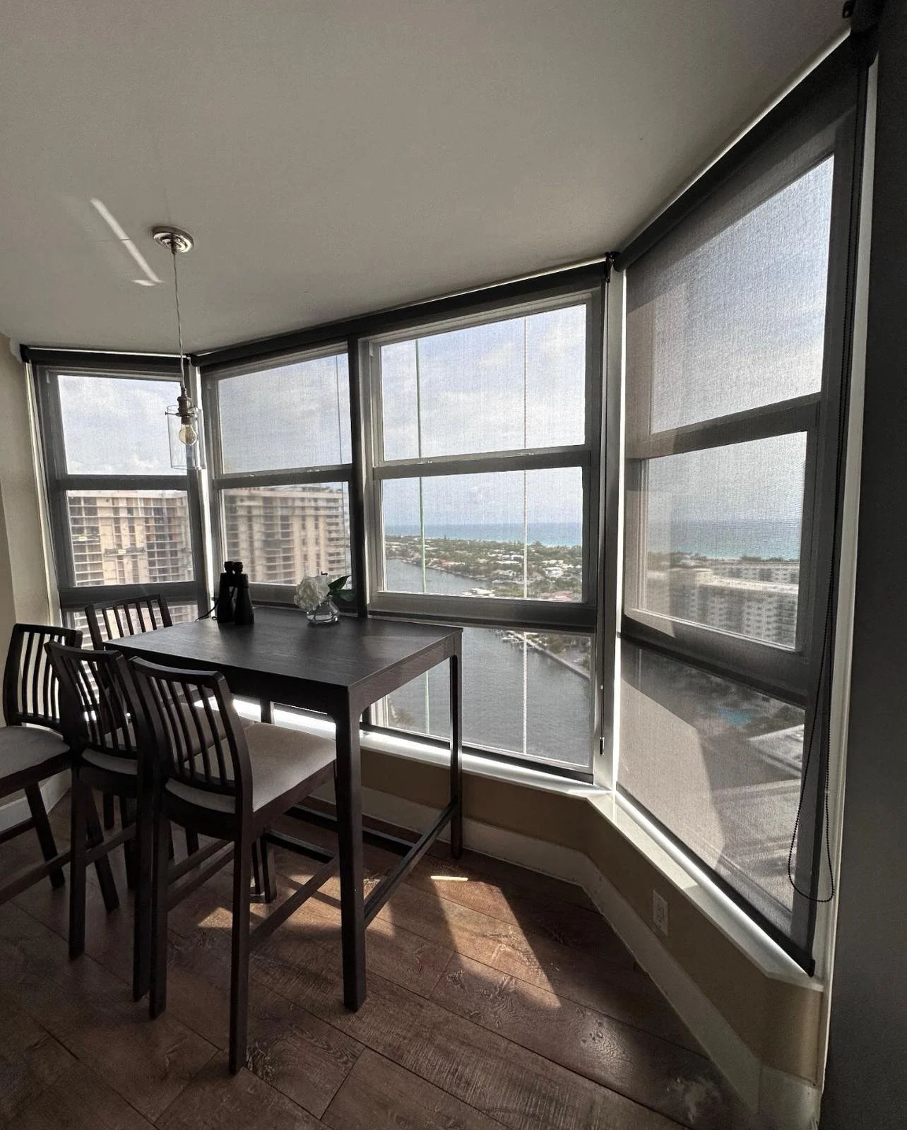Dining area with a dark wooden table and four matching chairs next to large windows with views of buildings, water, and sky outside.