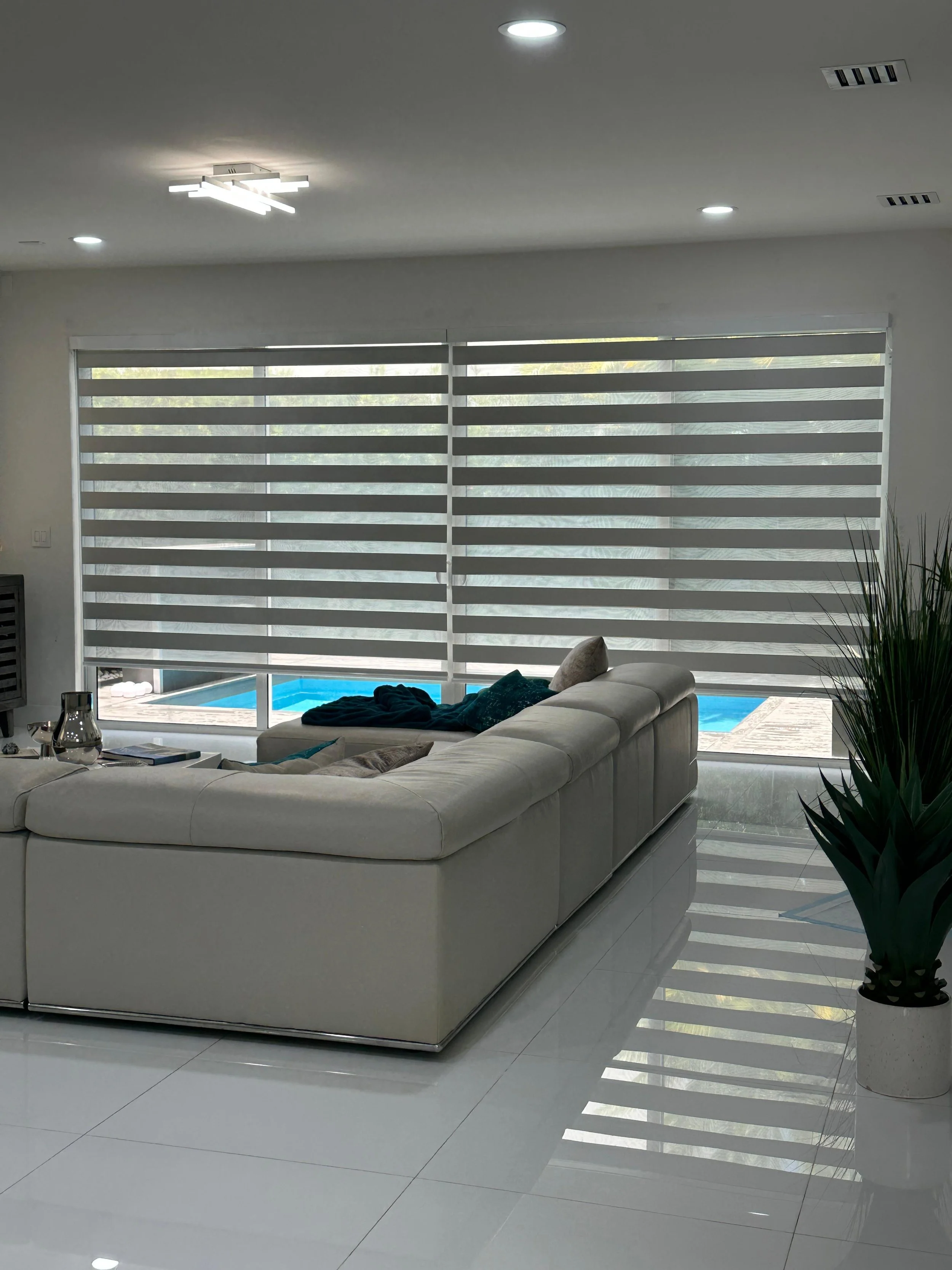 Modern living room with white sectional sofa, large window with horizontal blinds, potted plant, and view of a backyard pool.