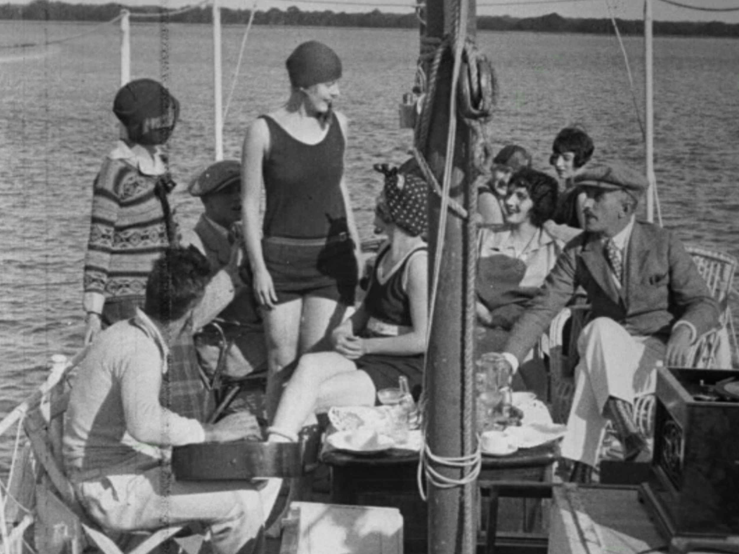 A group of people on a boat by a lake, dressed in vintage clothing, with some seated and others standing, engaging in conversation and enjoying the view.