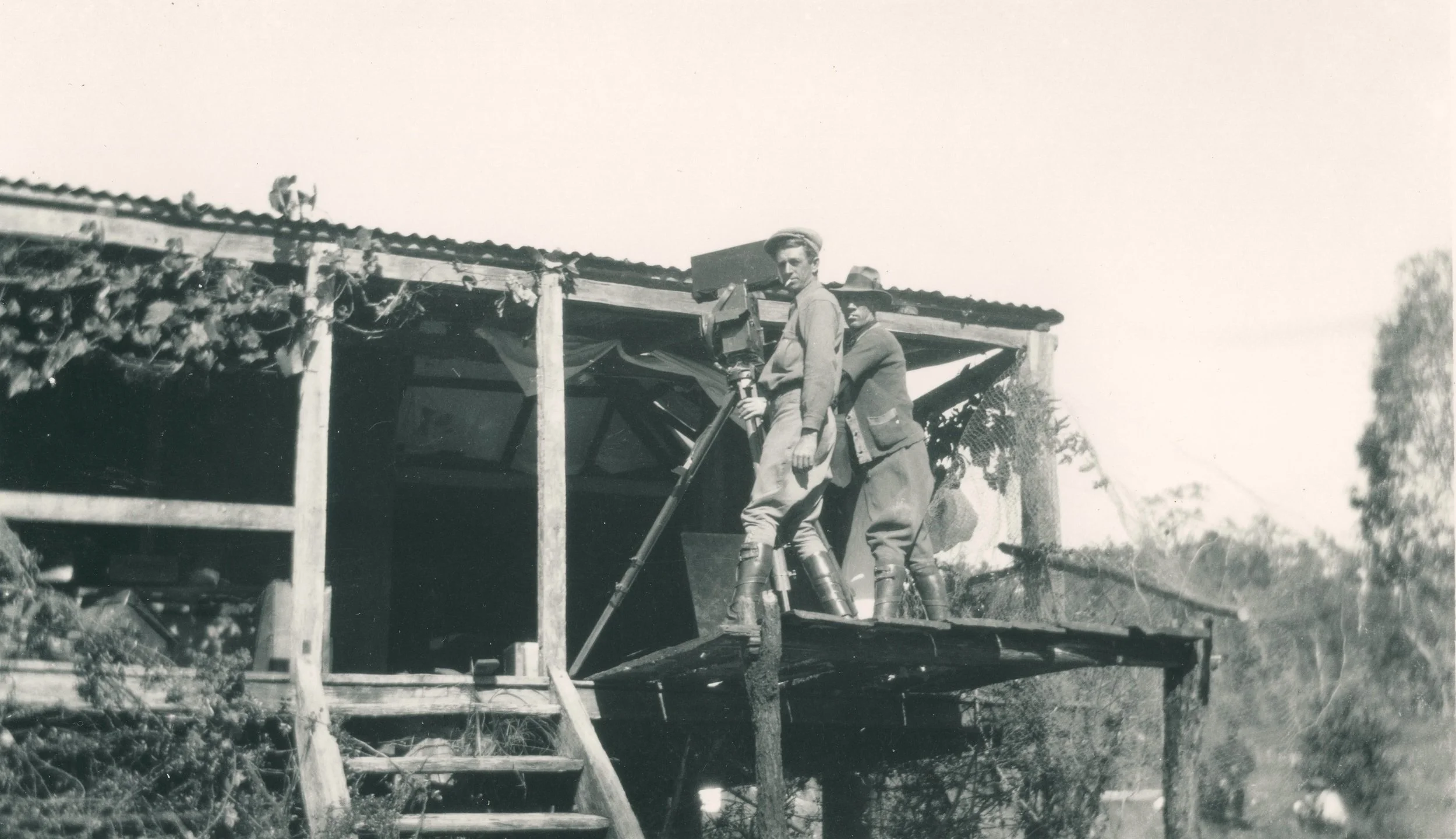 Black and white photo of two young men standing on a wooden platform, with one holding a camera, outside a rustic structure on stilts with a sloped roof, surrounded by trees.