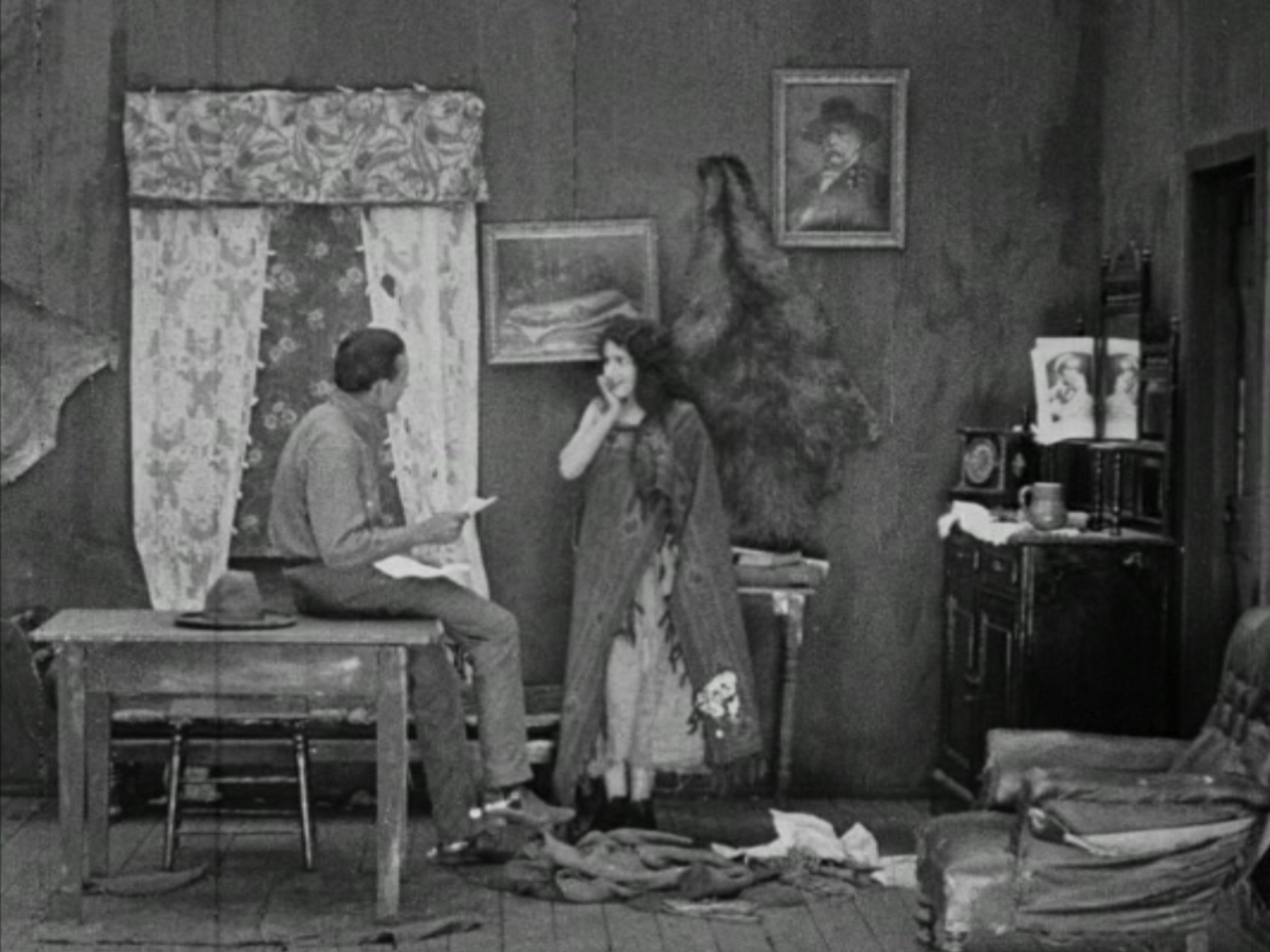 Interior scene with two women, one seated on a table and the other standing, in a room with clutter and framed pictures on the wall.