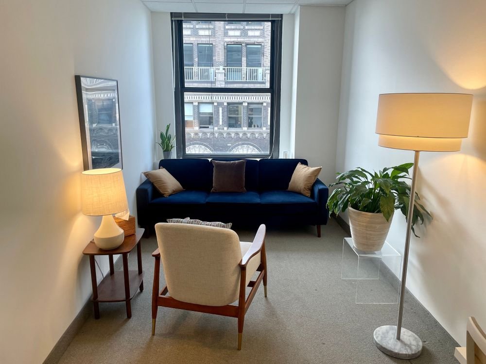 A cozy living room with a navy blue sofa, a cream armchair, a side table with a lamp, a tall floor lamp, a large potted plant, and a window with a city view.