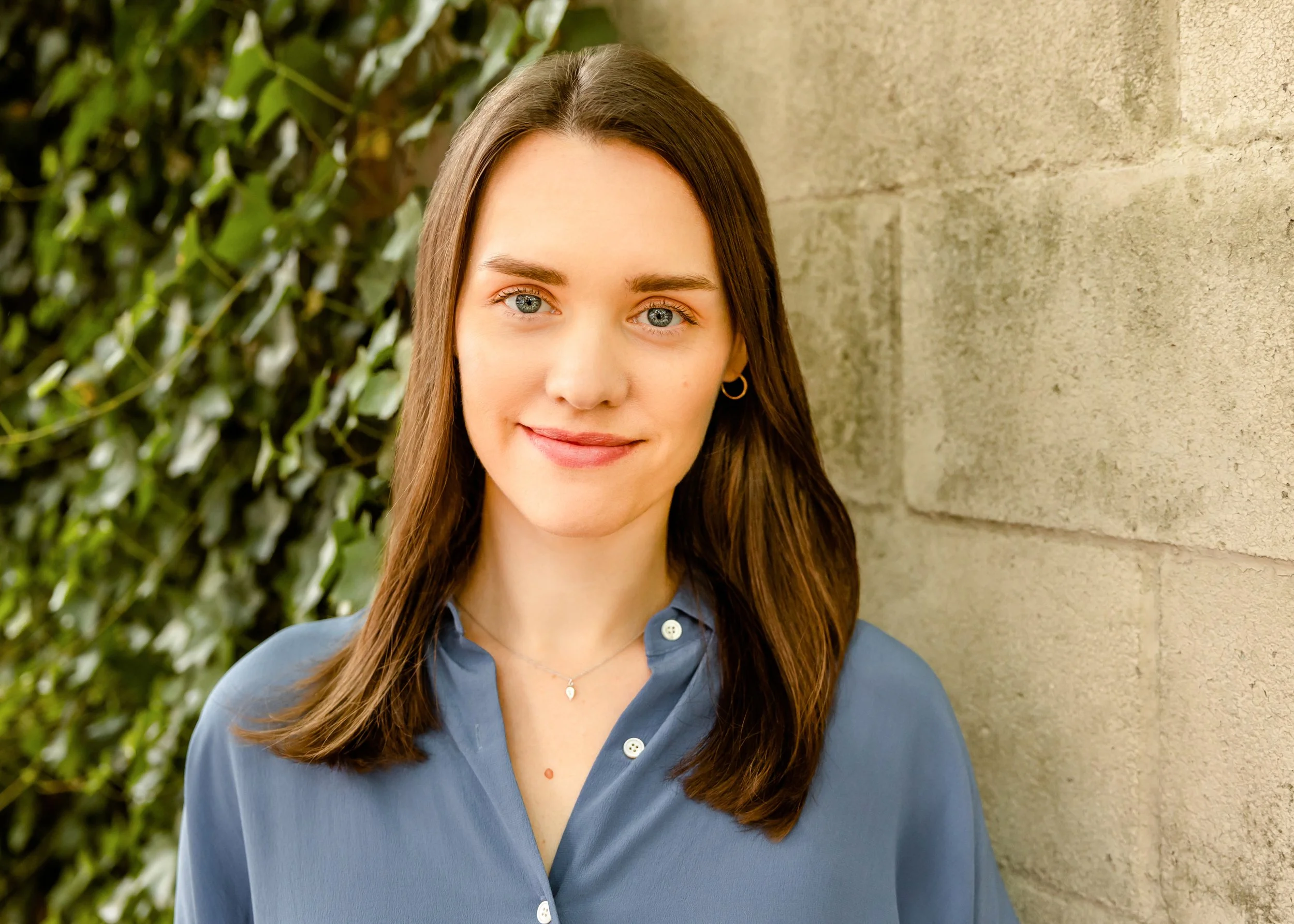A young woman with shoulder-length brown hair and blue eyes smiling outdoors beside a brick wall and green foliage.