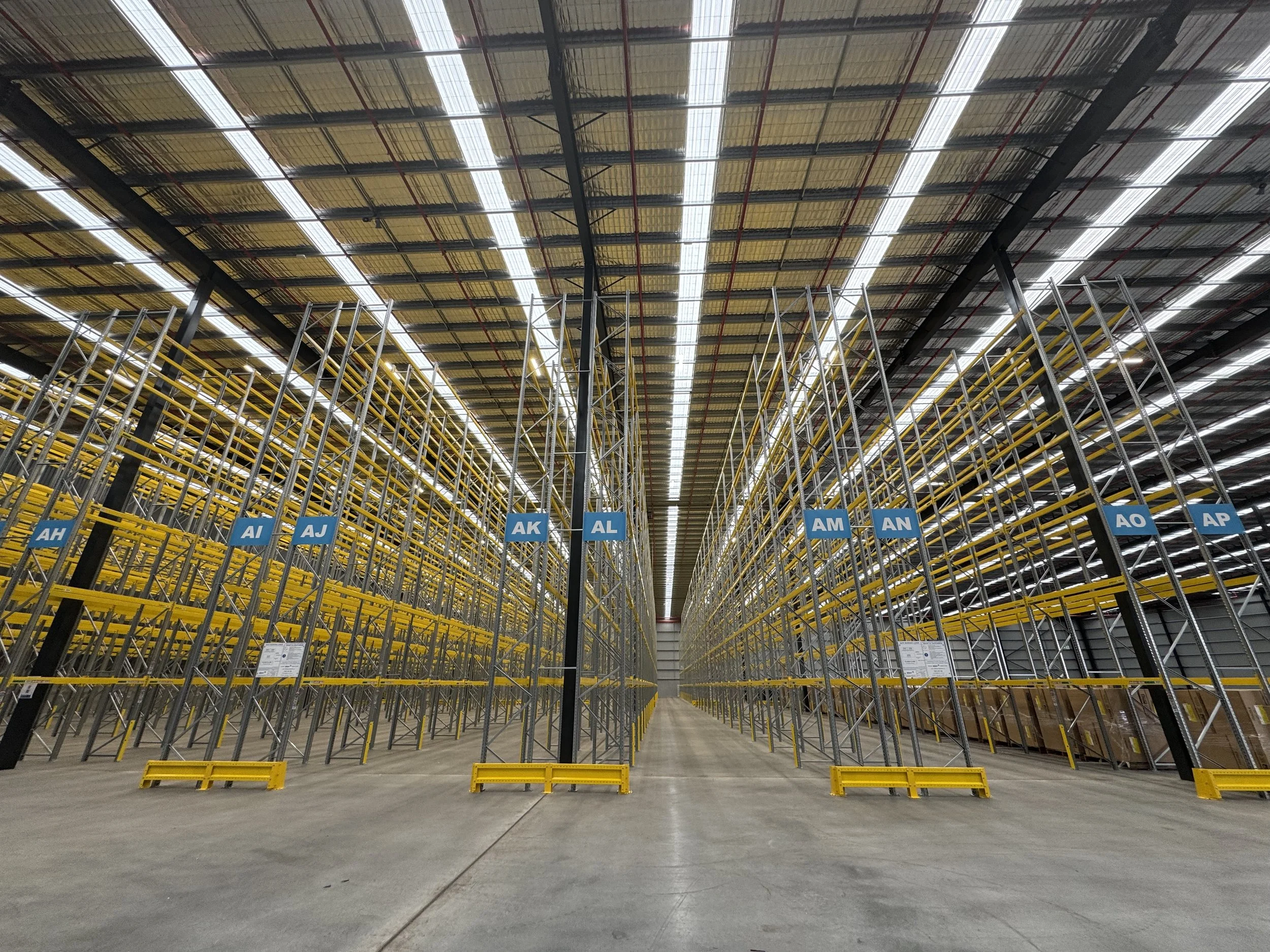 Empty warehouse aisle with tall metal shelves, yellow beams, and blue labels with letters and numbers, under a high ceiling with bright lighting.
