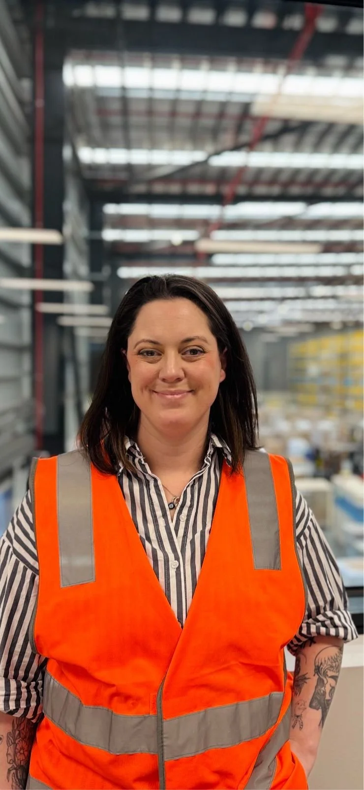 A woman wearing an orange safety vest and striped shirt standing in an industrial warehouse.