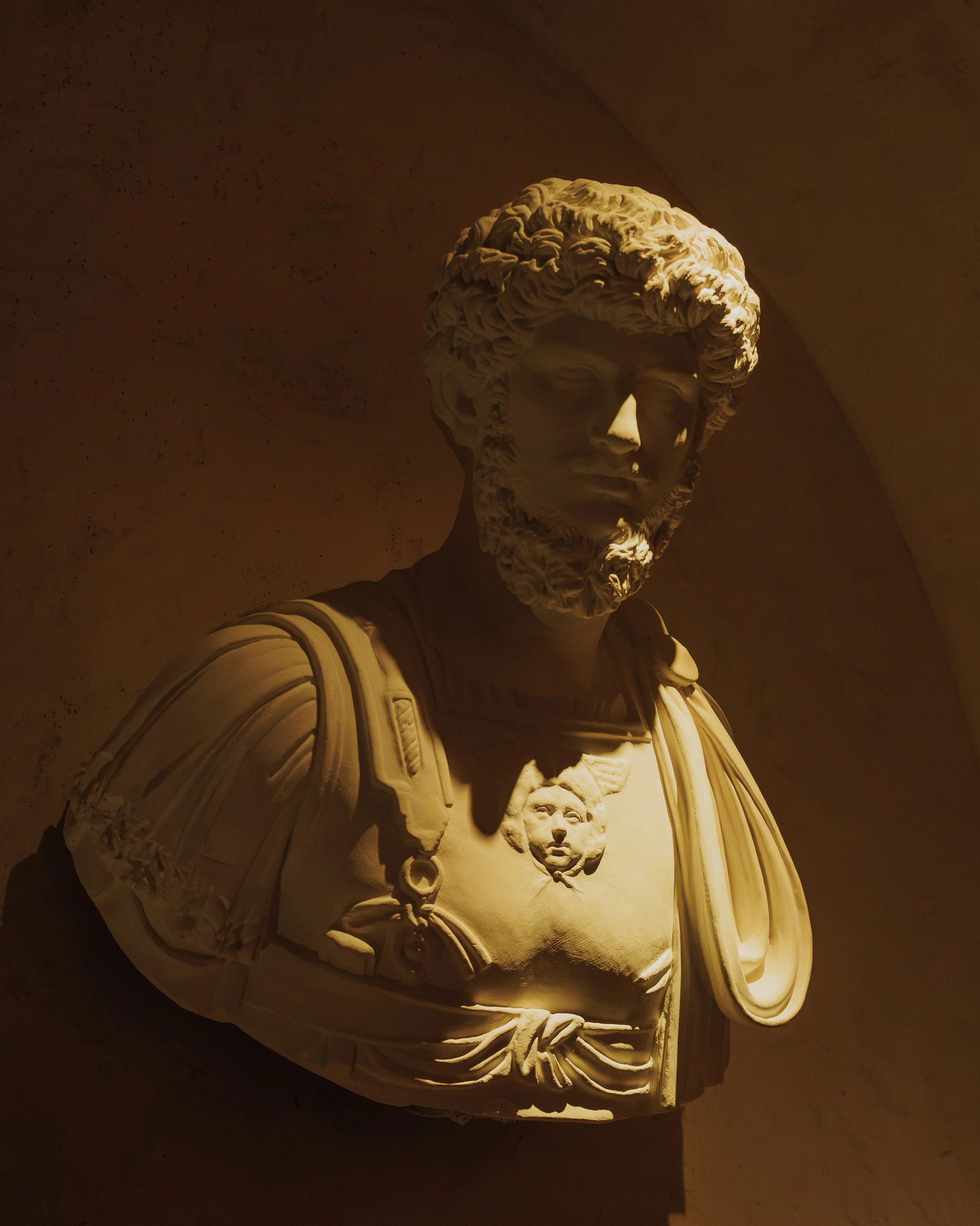 Close-up of an illuminated Roman bust wearing sculpted armor and drapery against a warm plaster wall.
