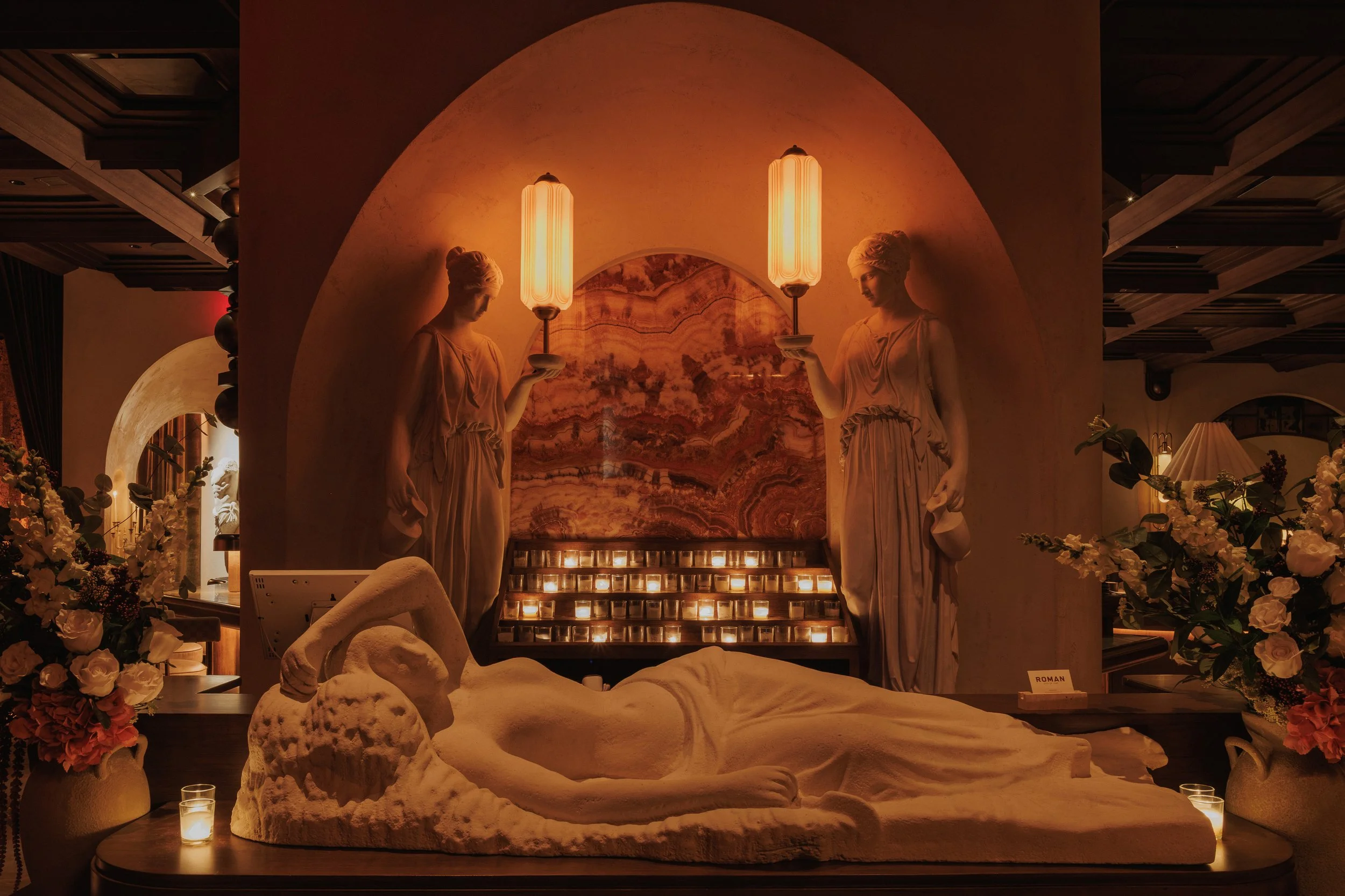 Reclining classical sculpture beneath a sculptural bottle display, floral arrangements and illuminated mural at Roman God of Fire.