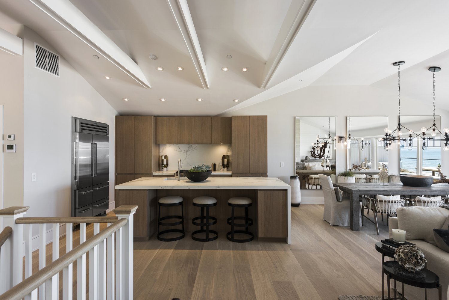 Open-concept living space with a modern kitchen island featuring a black bowl and three bar stools, adjacent to a dining area with a dark wooden table and white chairs, and a living room with a white sofa, decorative items, and large windows overlook