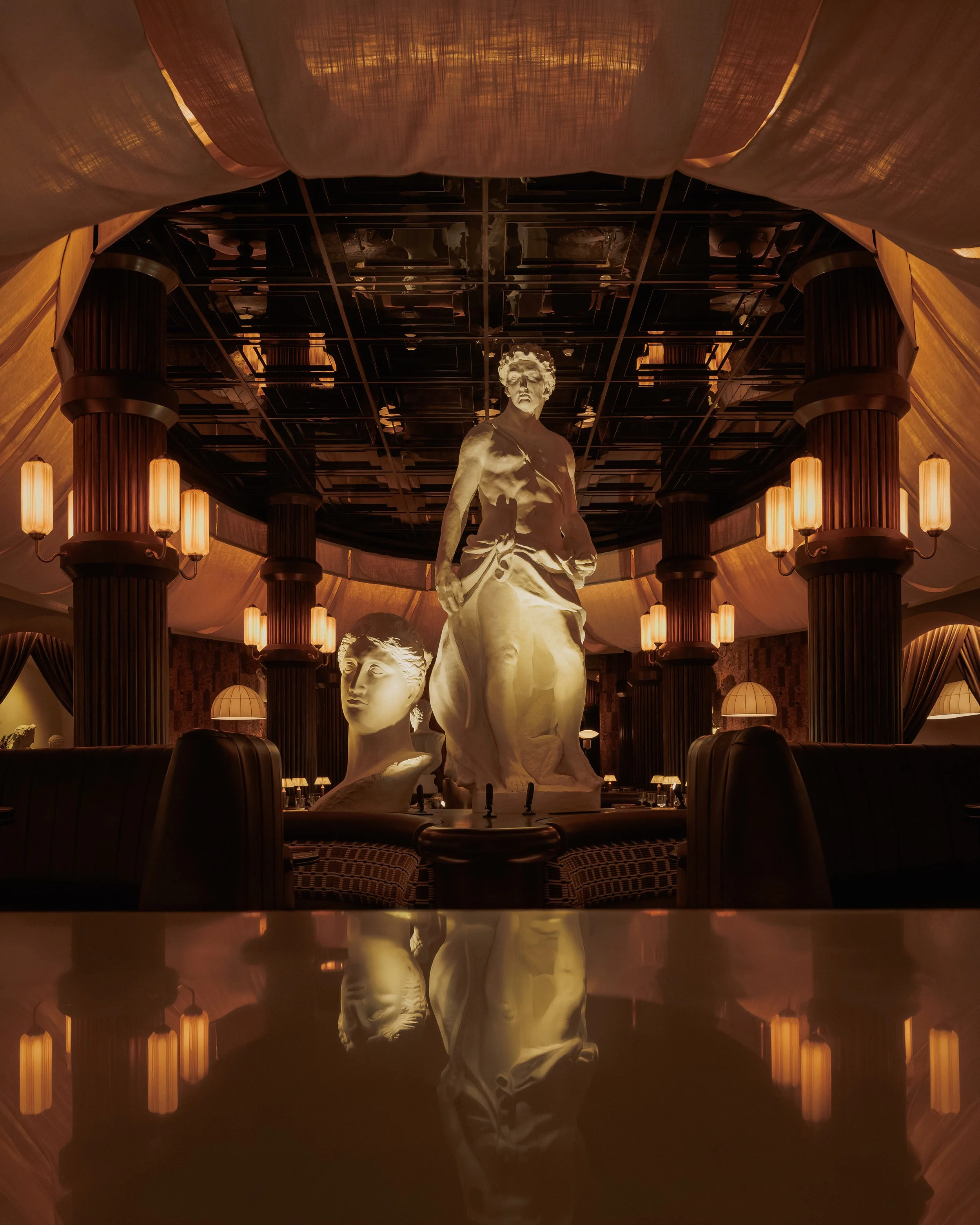 Long view through the dining room toward monumental classical sculptures, dark columns and a reflective foreground surface.