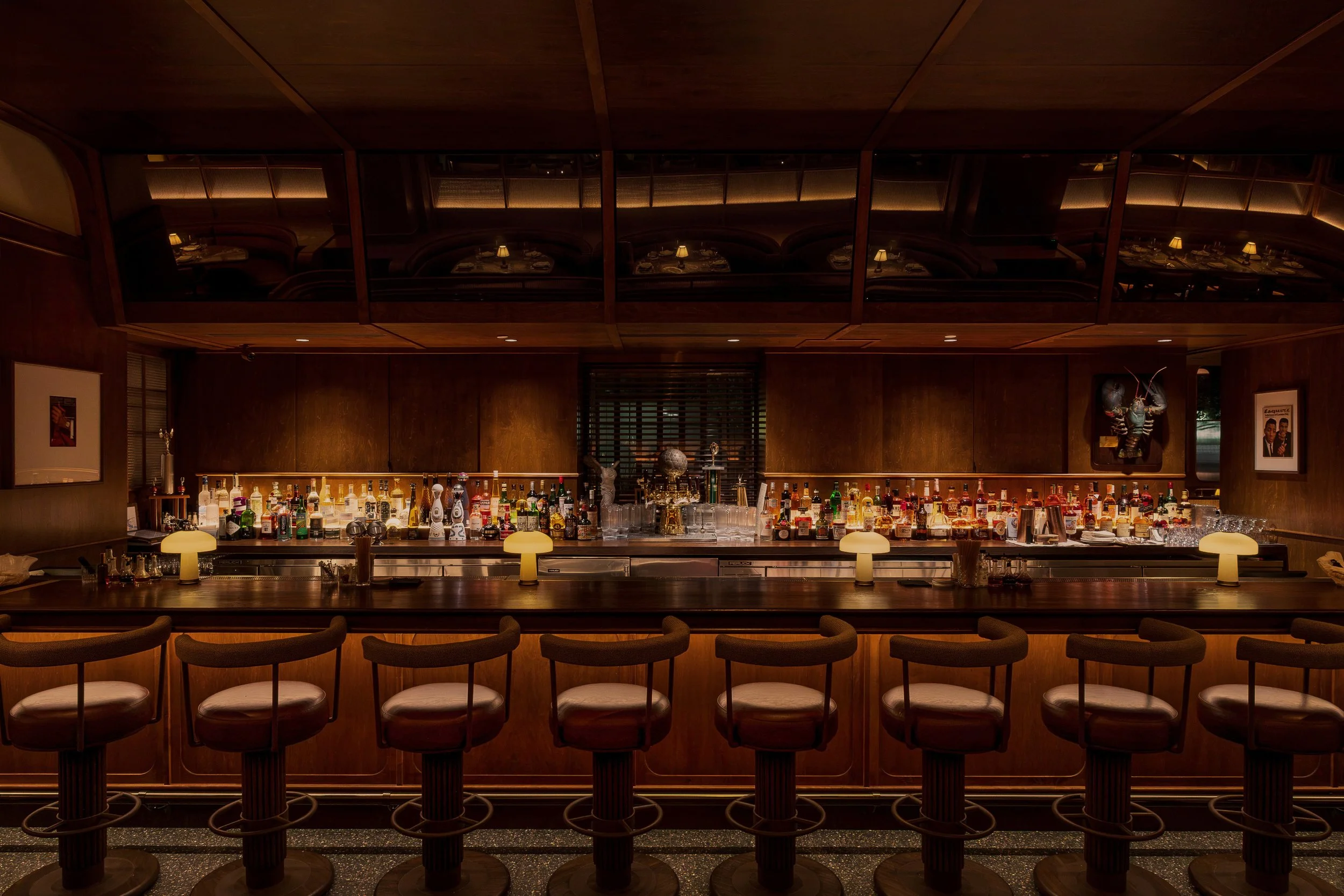 The central bar combines illuminated bottle displays, dark wood millwork and tailored counter seating beneath a reflective overhead canopy.