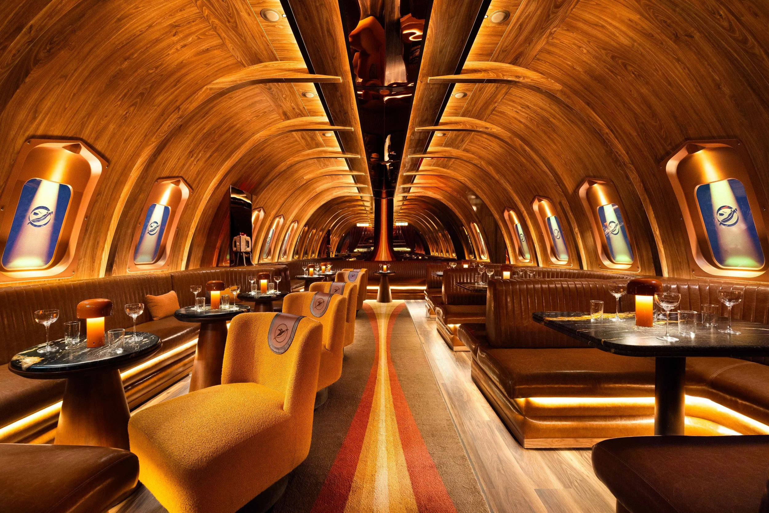 Interior of Carry On Airlines in Phoenix, designed by Peter Bowden and PETERMAX Co. as an immersive 1970s luxury jetliner, with curved leather banquettes, illuminated aircraft windows, intimate cocktail tables and warm amber lighting.