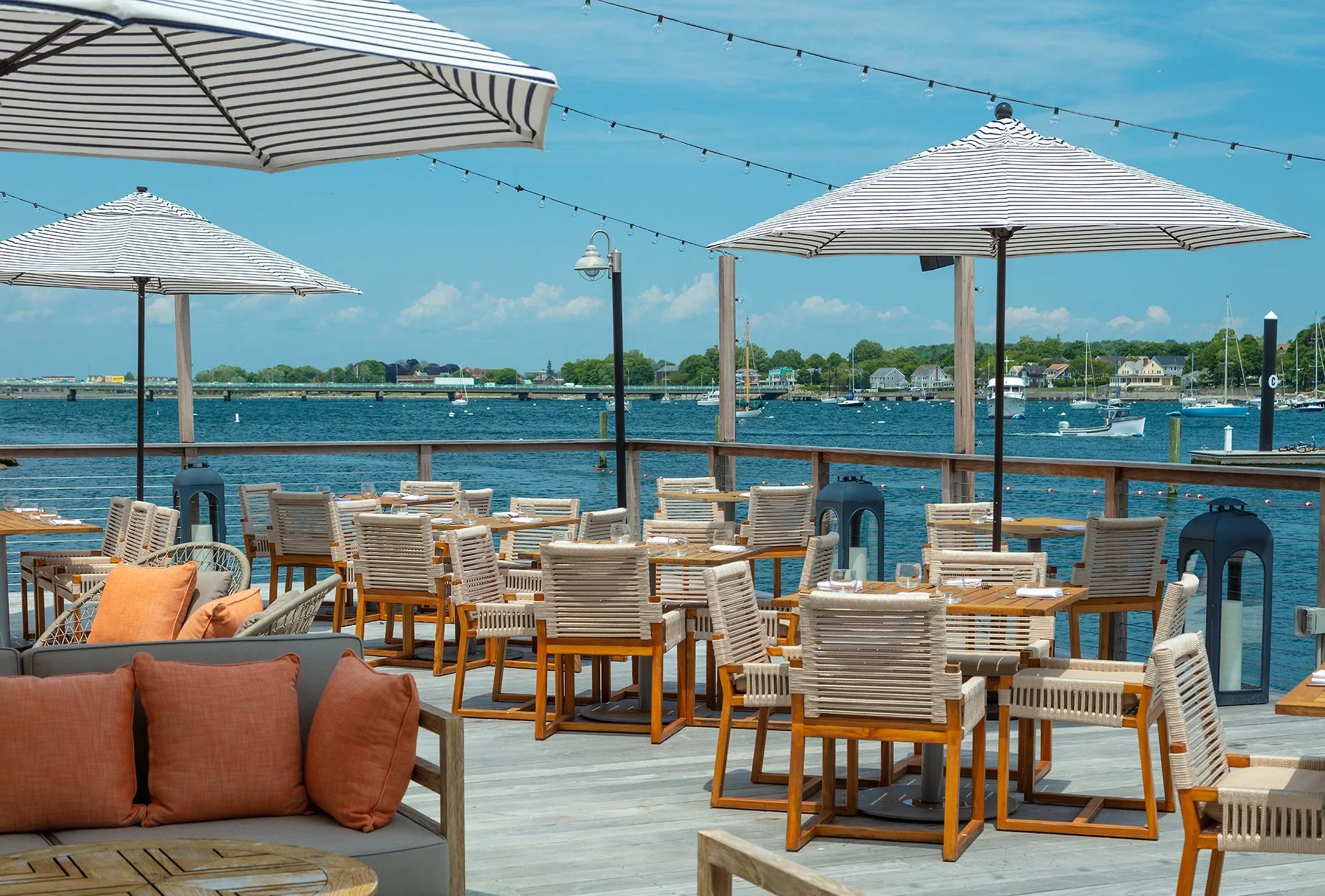 Gurney’s Newport on the nautical deck of Scarpetta restaurant designed by Peter Maxwell Bowden of PETERMAX Co., featuring rope-wrapped lounge chairs, honeyed teak wood, striped umbrellas, and beautiful harbor views.