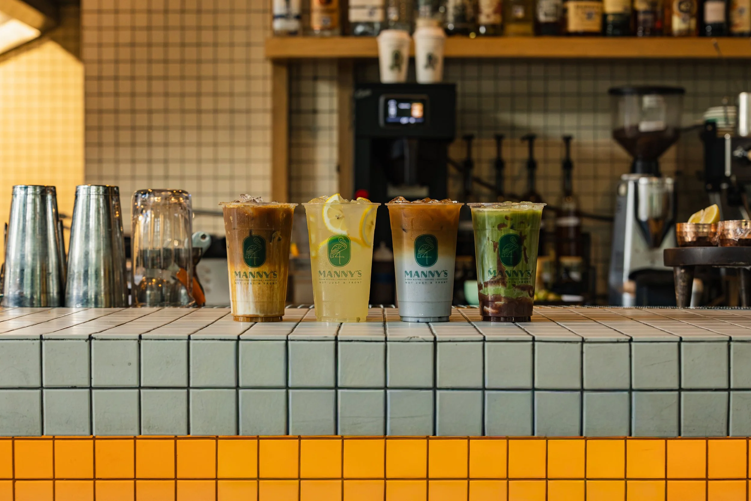 Four specialty drinks in branded cups on the blue-tiled bar at Manny’s, with a glowing undercounter lighting.