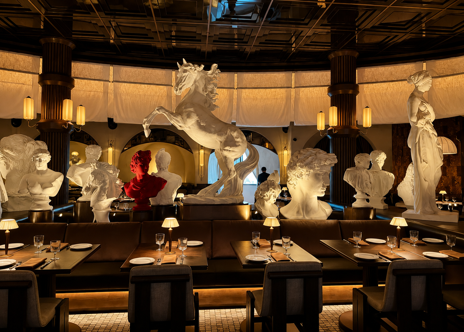 Dining room at Roman God of Fire in Tempe, featuring a gallery of illuminated classical sculptures, a rearing horse, curved leather banquettes and amber table lamps beneath a dark coffered ceiling.