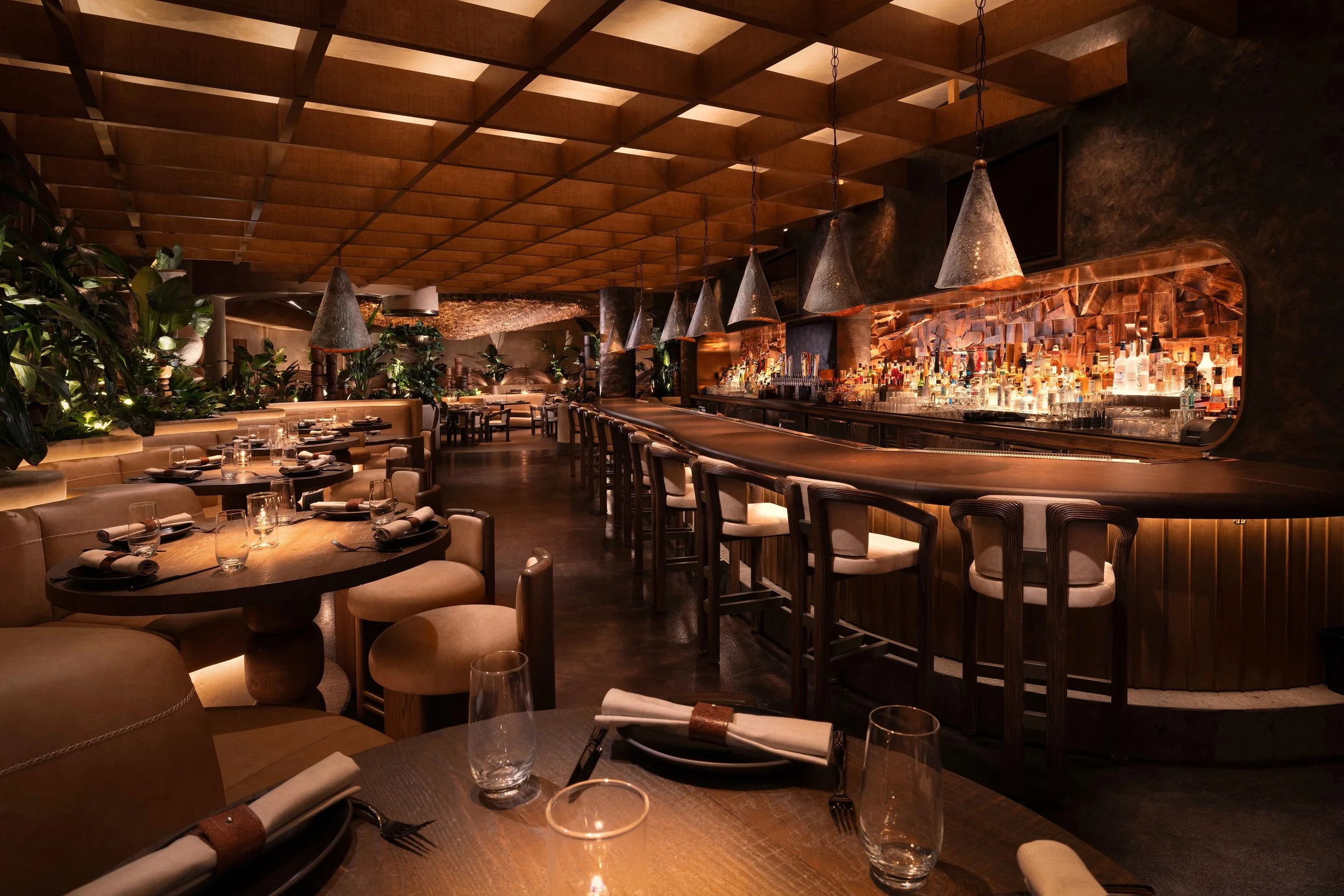 Amaya Modern Mexican dining room at The Cosmopolitan of Las Vegas, featuring a curved banquette, tropical greenery, woven dining chairs and a dramatic sculptural wood wall evocative of Equatorial Brutalism.
