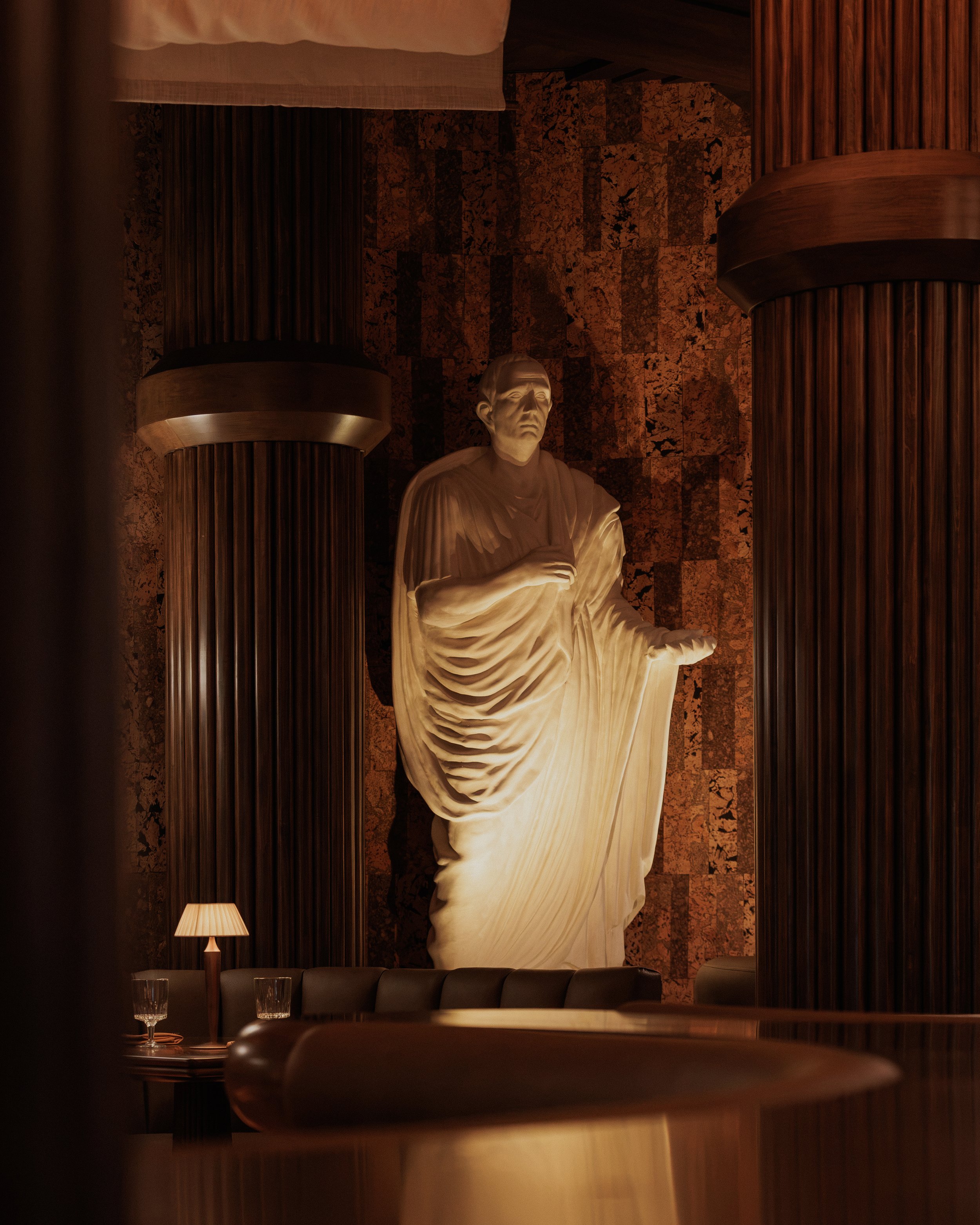Draped classical statue framed by dark fluted columns and a warm metallic wall inside the Tempe restaurant.