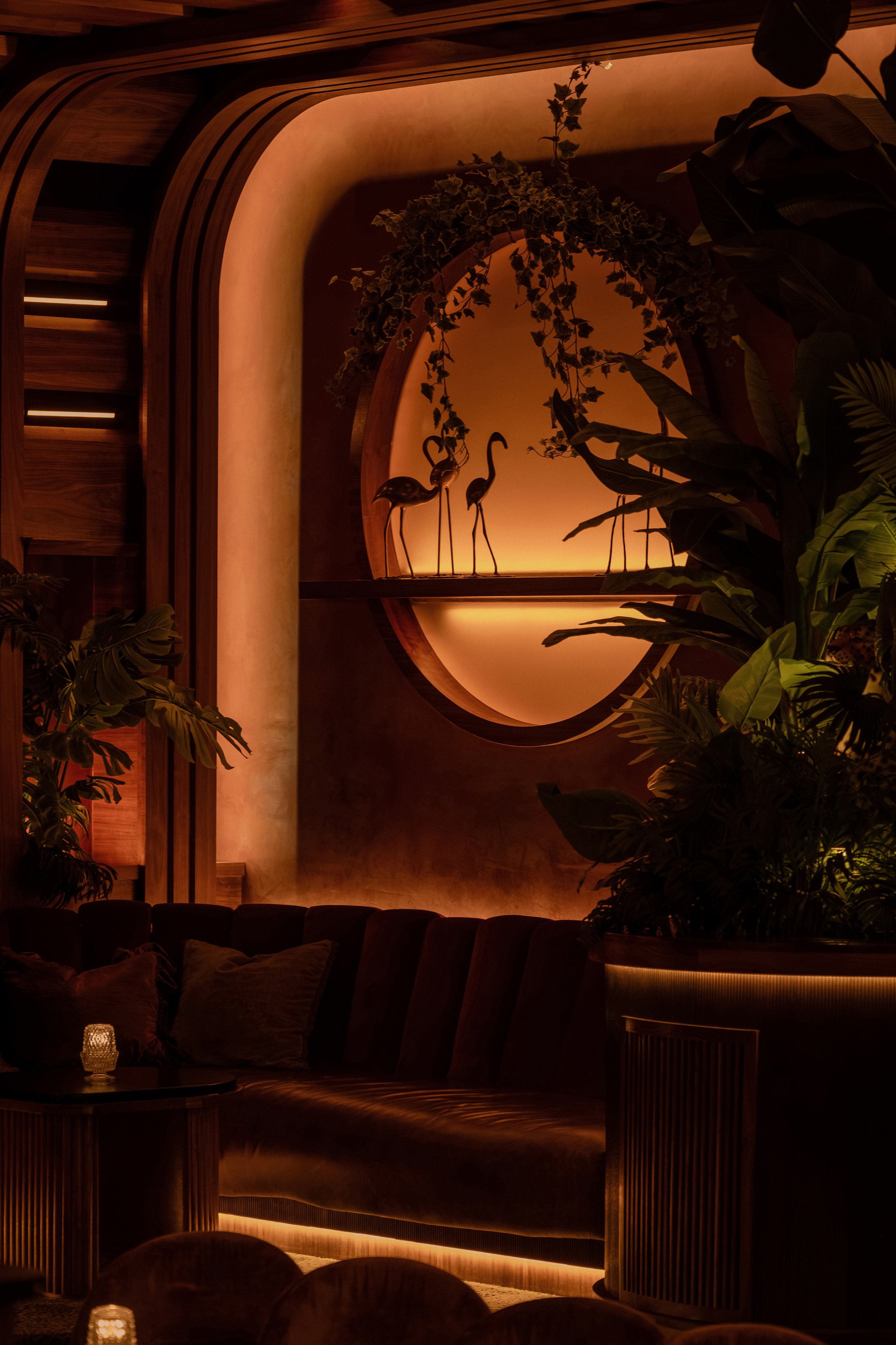 Intimate curved booth at Powder Room with dark upholstery, tropical foliage and a softly illuminated wall mirror.