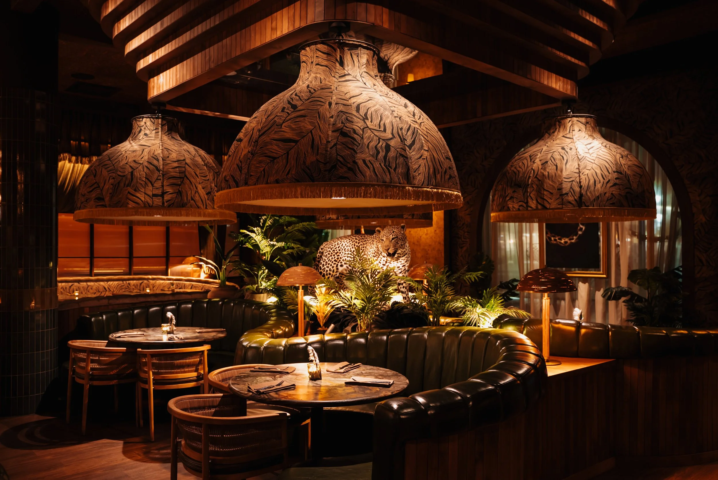 Interior of Filthy Animal in Tempe, featuring an olive-green leather banquette, woven wood chairs, glossy green tile, tropical planting and dramatic animal imagery.