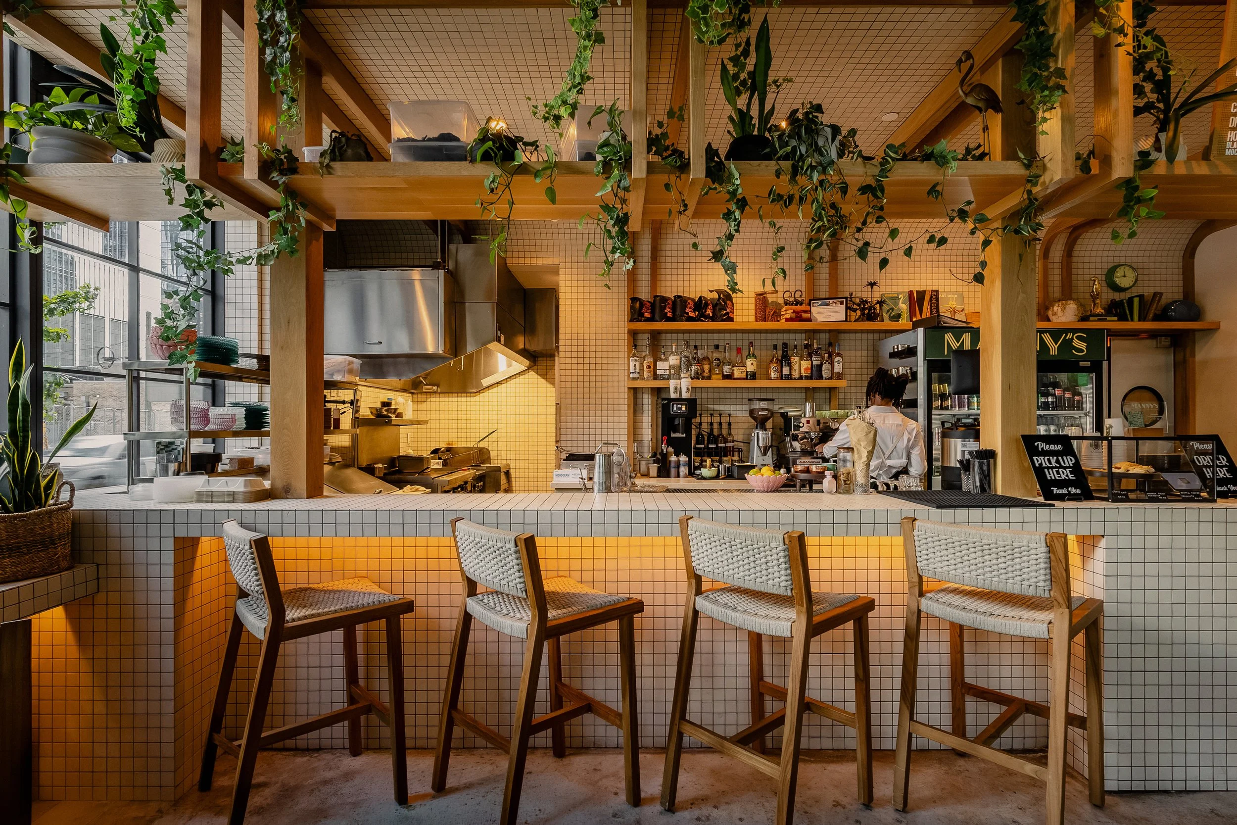 Front view of the Manny’s bar with woven stools, white tile, warm wood finishes and hanging plants.