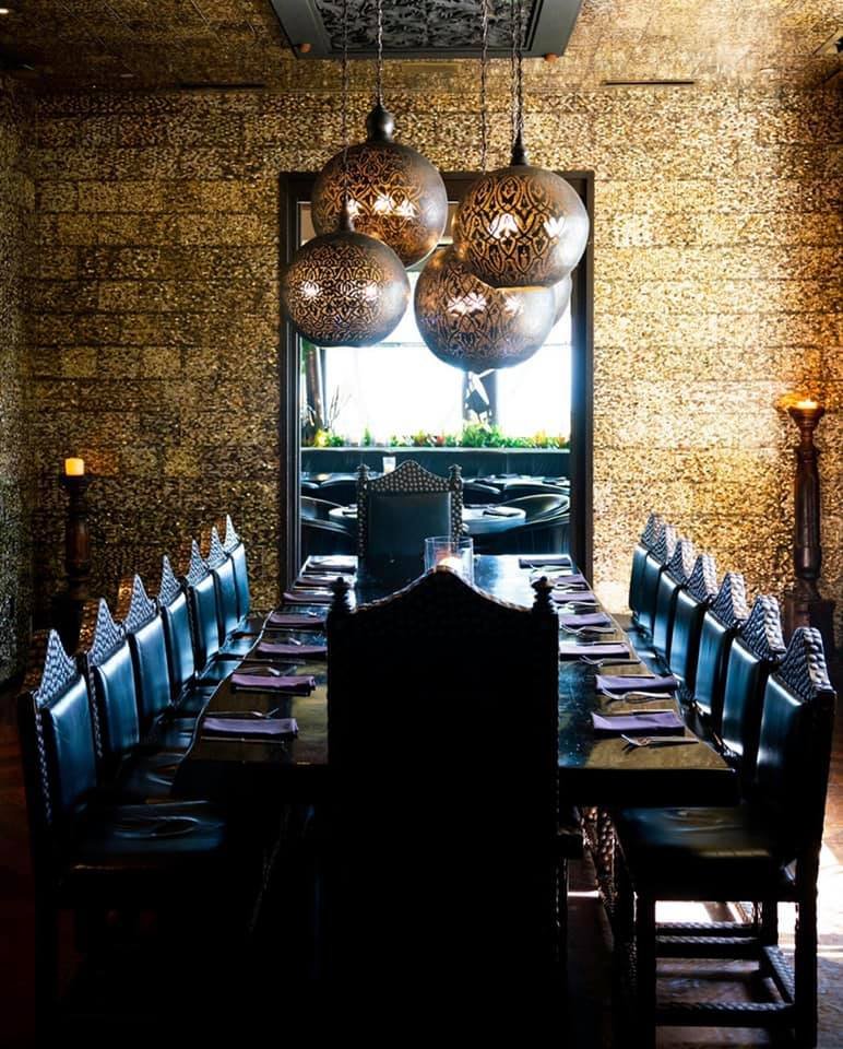 A dimly lit restaurant dining room with a long table set with napkins and glasses. Four pendant lights with intricate patterns hang overhead, and there is a window at the end of the room letting in natural light. The walls are textured and decorative