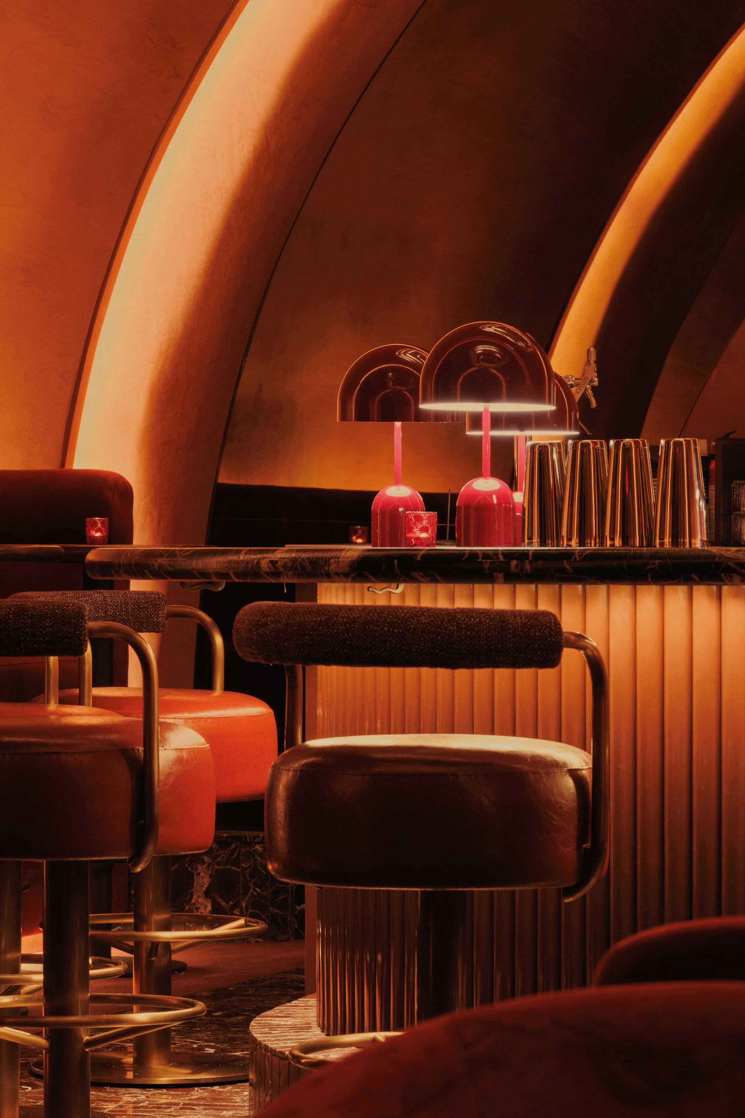 Low-angle view between upholstered lounge chairs toward Mea Culpa’s glowing arches, layered chandeliers and dramatic central bar.