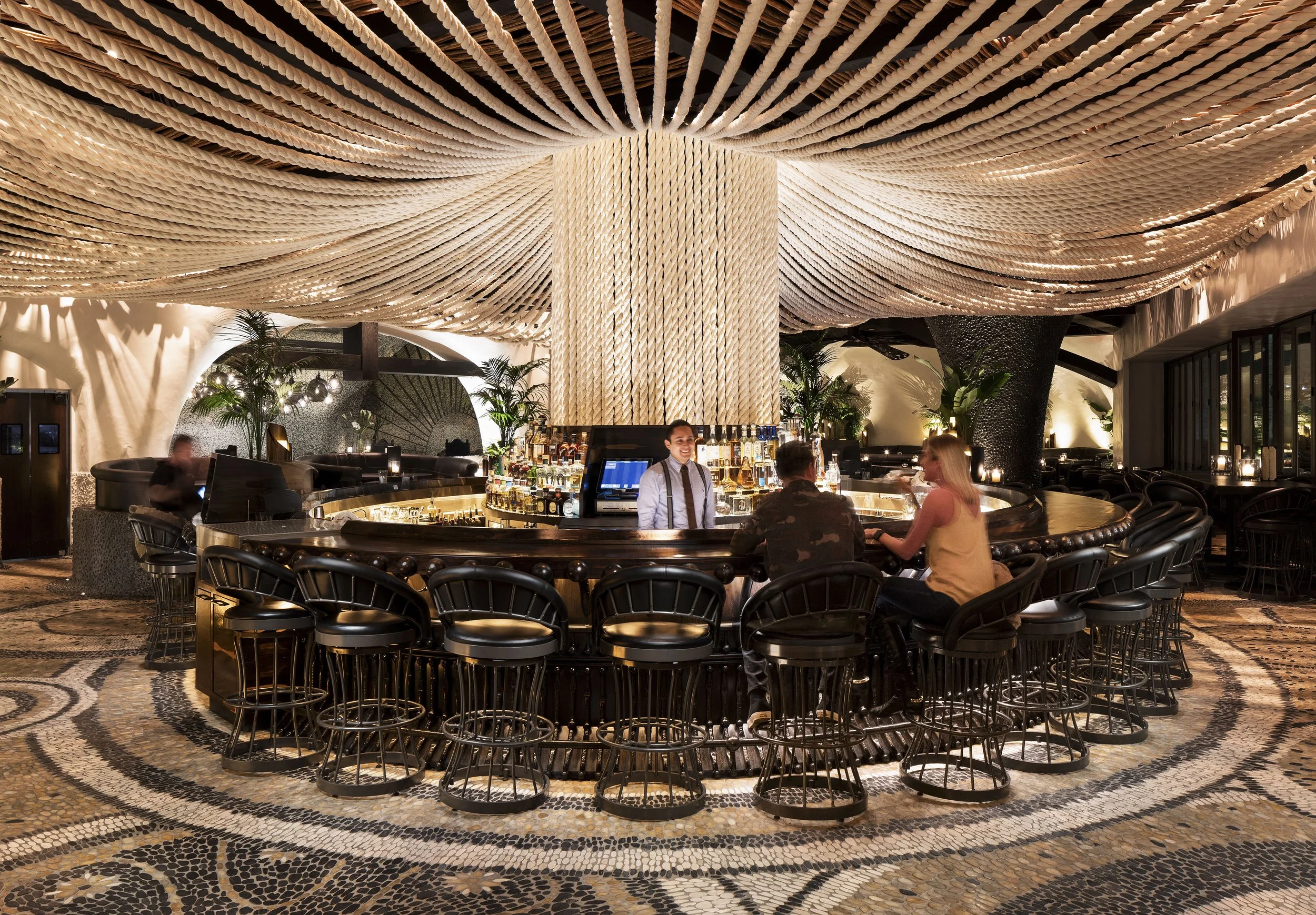 Bar interior at Javier’s La Jolla, featuring a sculptural rope installation across the ceiling, a rope-wrapped column, dark leather barstools and hand-laid mosaic flooring.