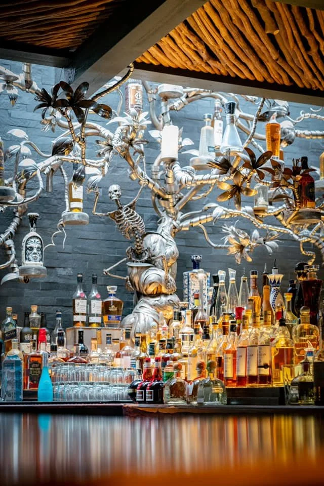 Bar with a silver decorative tree structure holding bottles and skull decor, bottles of alcohol on the counter, and a modern interior with a wooden ceiling.