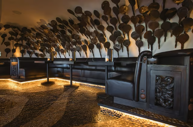 Decorative wall featuring numerous black and gold spoons, with black leather seating and warm ambient lighting in a restaurant or lounge.