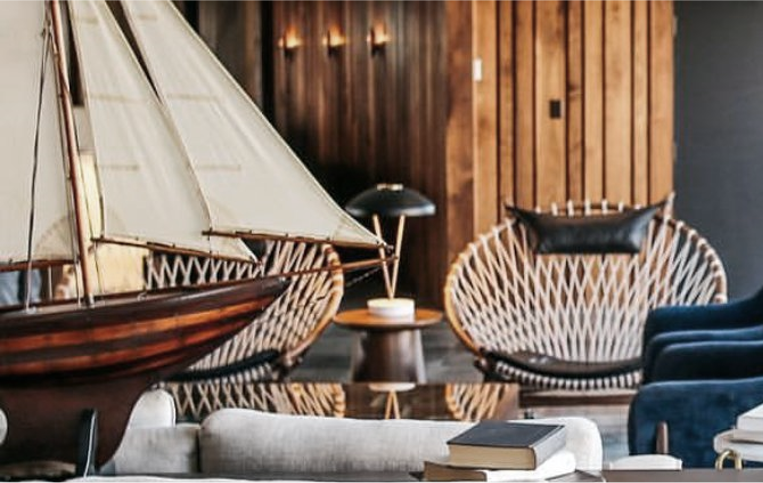 Living room with nautical theme, featuring a model ship with sails, a wooden side table, modern wicker chairs, a dark table lamp, and a dark wood-paneled wall.