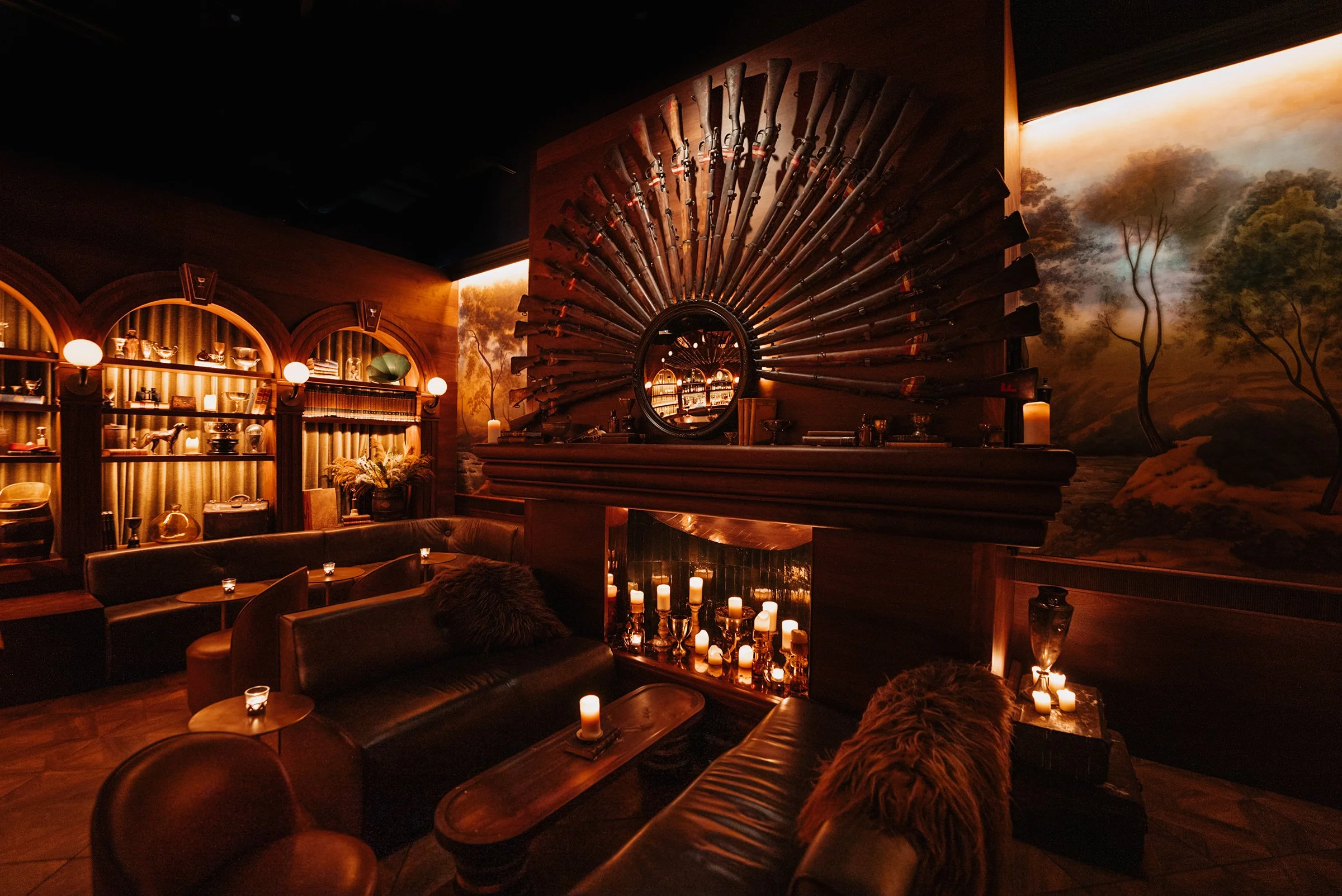Trophy Room in Phoenix showing a fireplace seating area surrounded by an arrangement of hunting rifles above the hearth, dark wood slat walls, green velvet drapery and warm globe lighting.