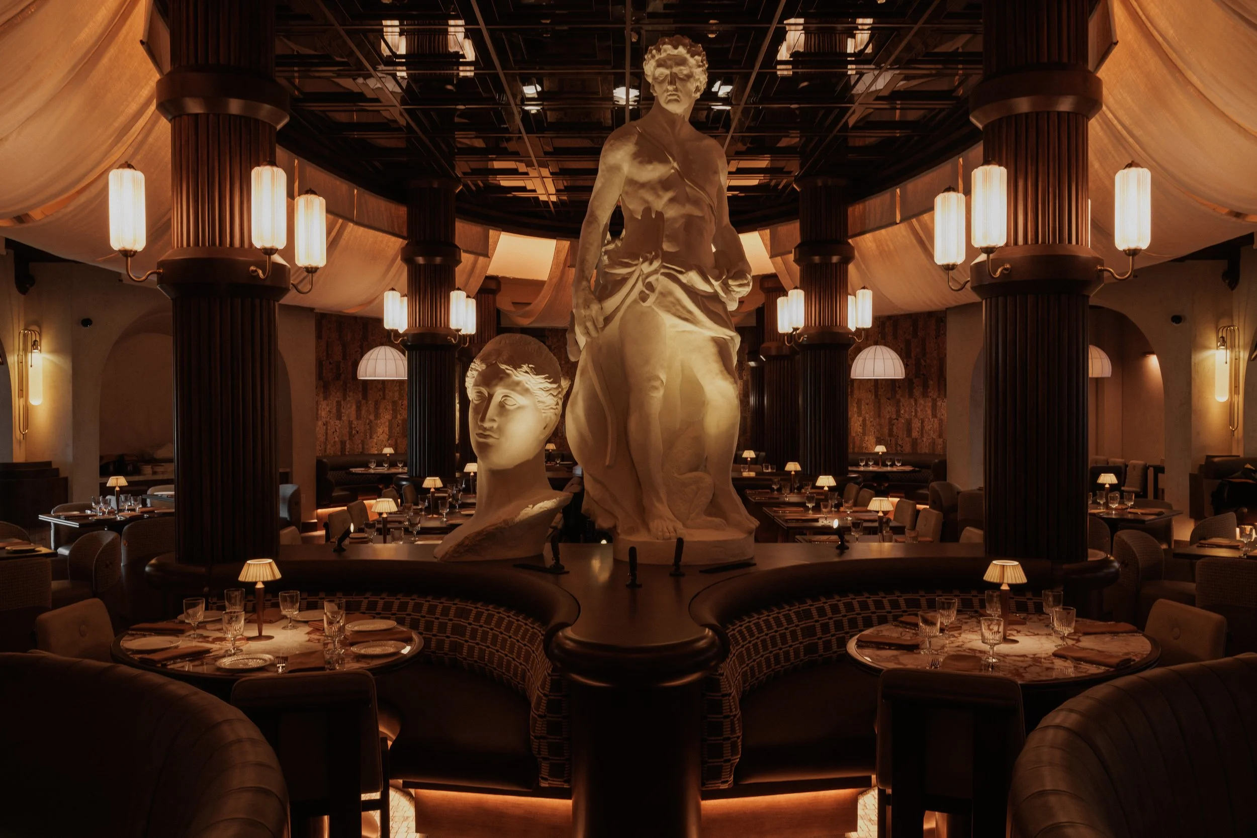 Symmetrical view of Roman God of Fire’s dining room, centered on monumental classical sculptures surrounded by curved booths and dark fluted columns.