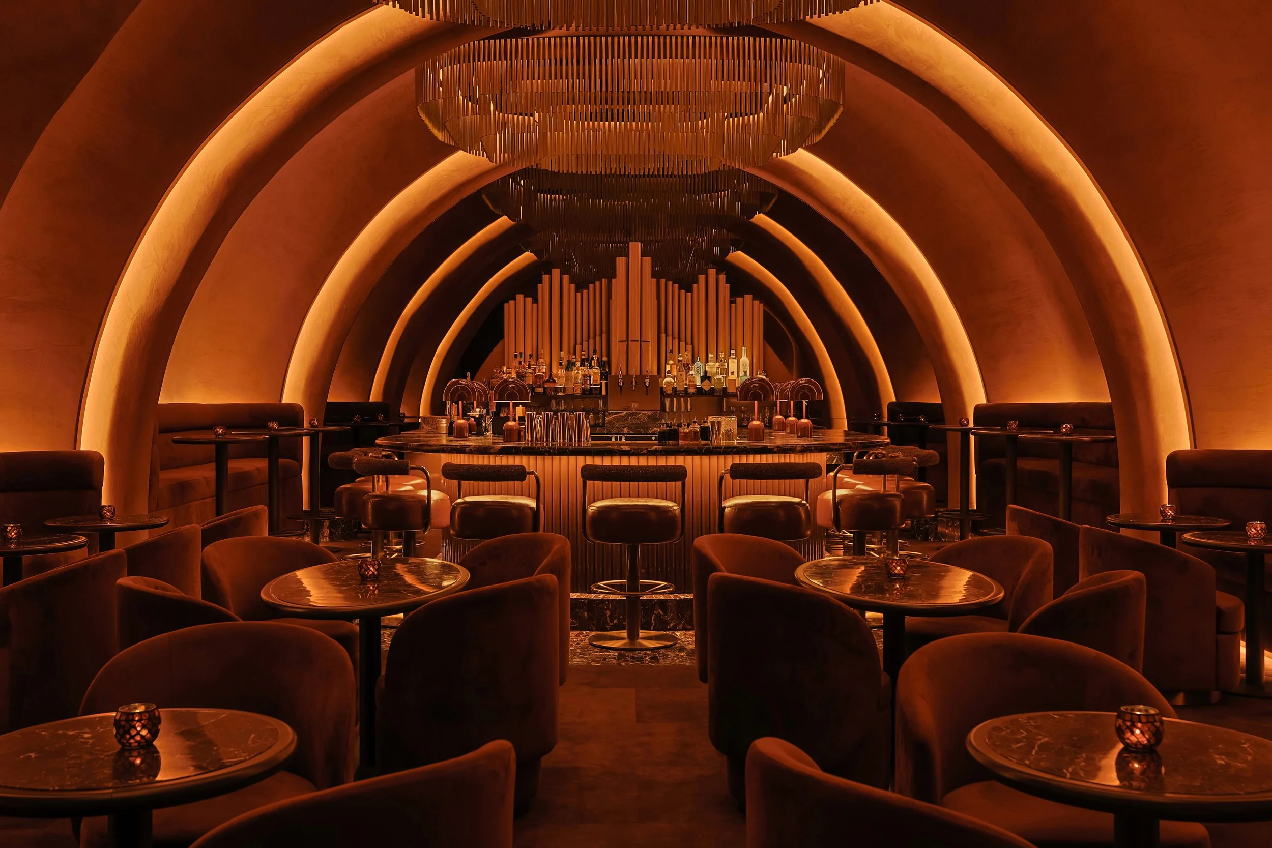 Symmetrical view through Mea Culpa’s intimate lounge seating toward the organ-inspired bar, framed by glowing arches and layered metal chandeliers.
