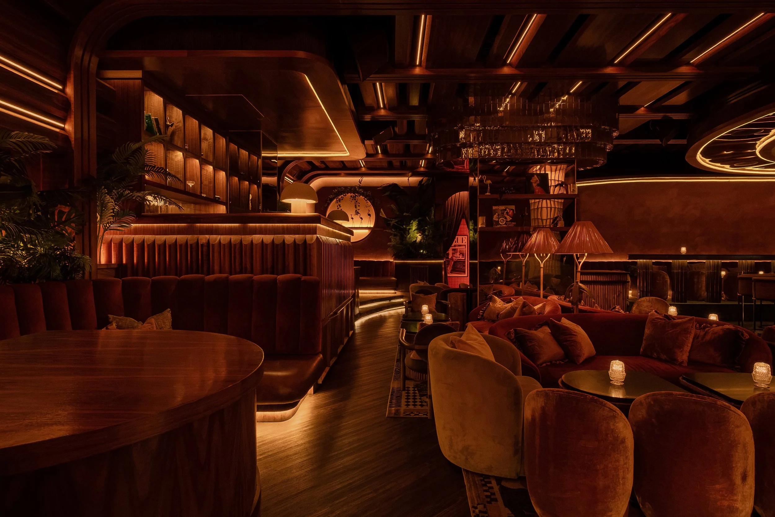 Curved banquette and velvet lounge chairs at Powder Room, framed by warm wood paneling and amber accent lighting.