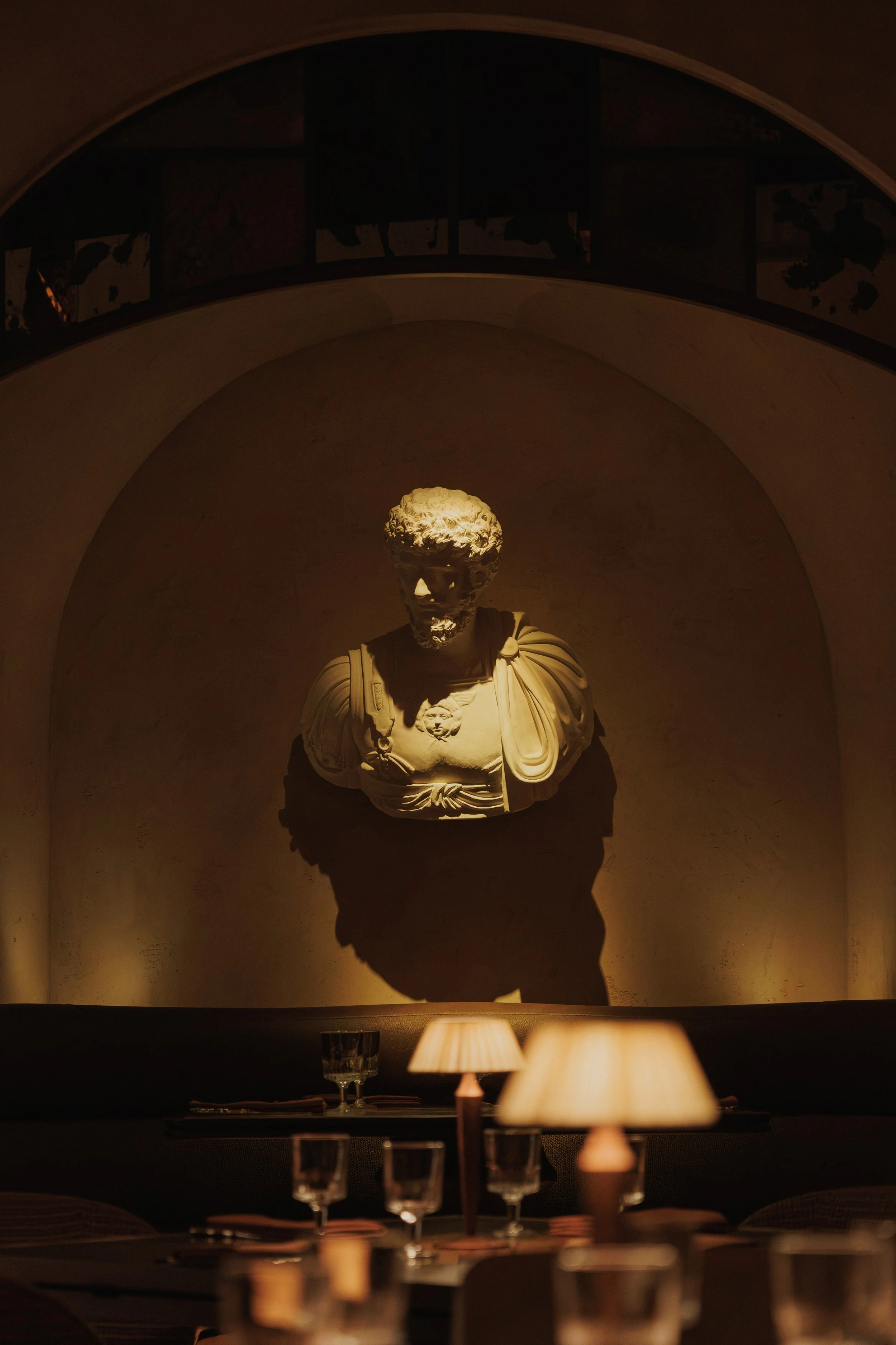 Classical bust centered within a softly illuminated arched niche above intimate candlelit dining tables at Roman God of Fire.