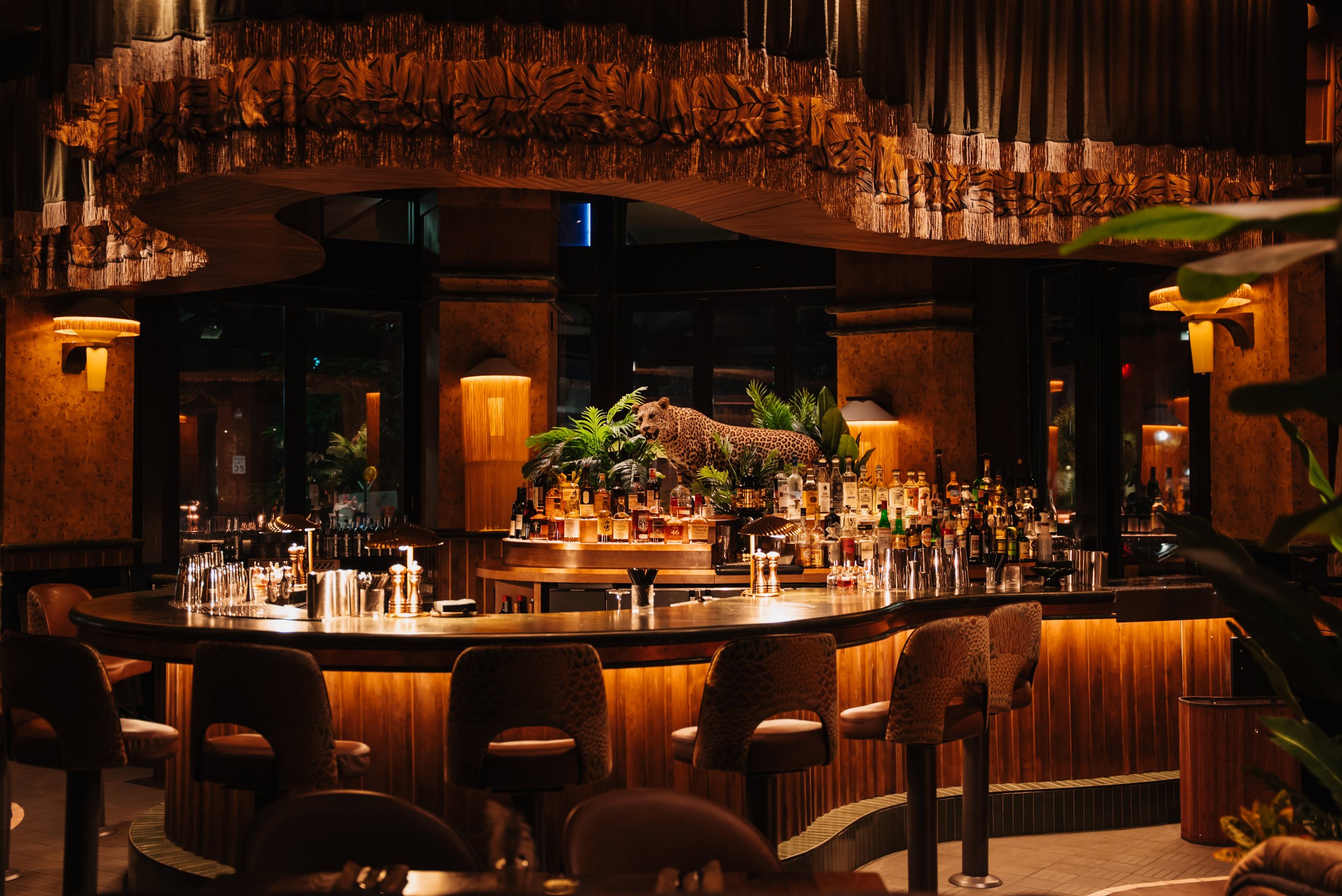 A lush, 1970s-inspired restaurant interior designed for Pretty Decent Concepts by the PETERMAX Co design team, featuring green banquettes, tropical planting, wood paneling, sculptural pendant lights, and patterned furnishings.