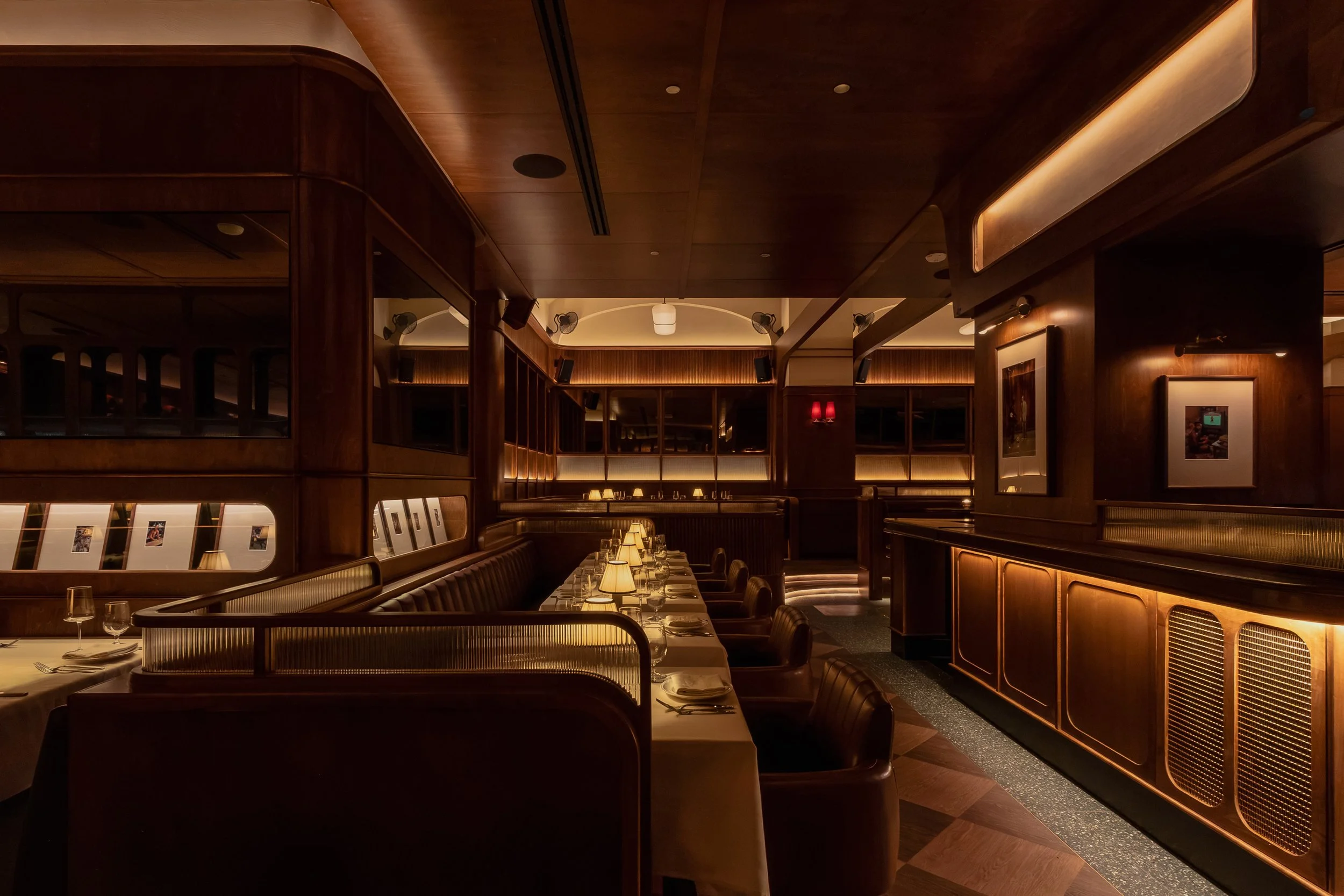 A linear view through the club reveals layered banquettes, marble-topped tables, illuminated shelving and dark wood millwork.